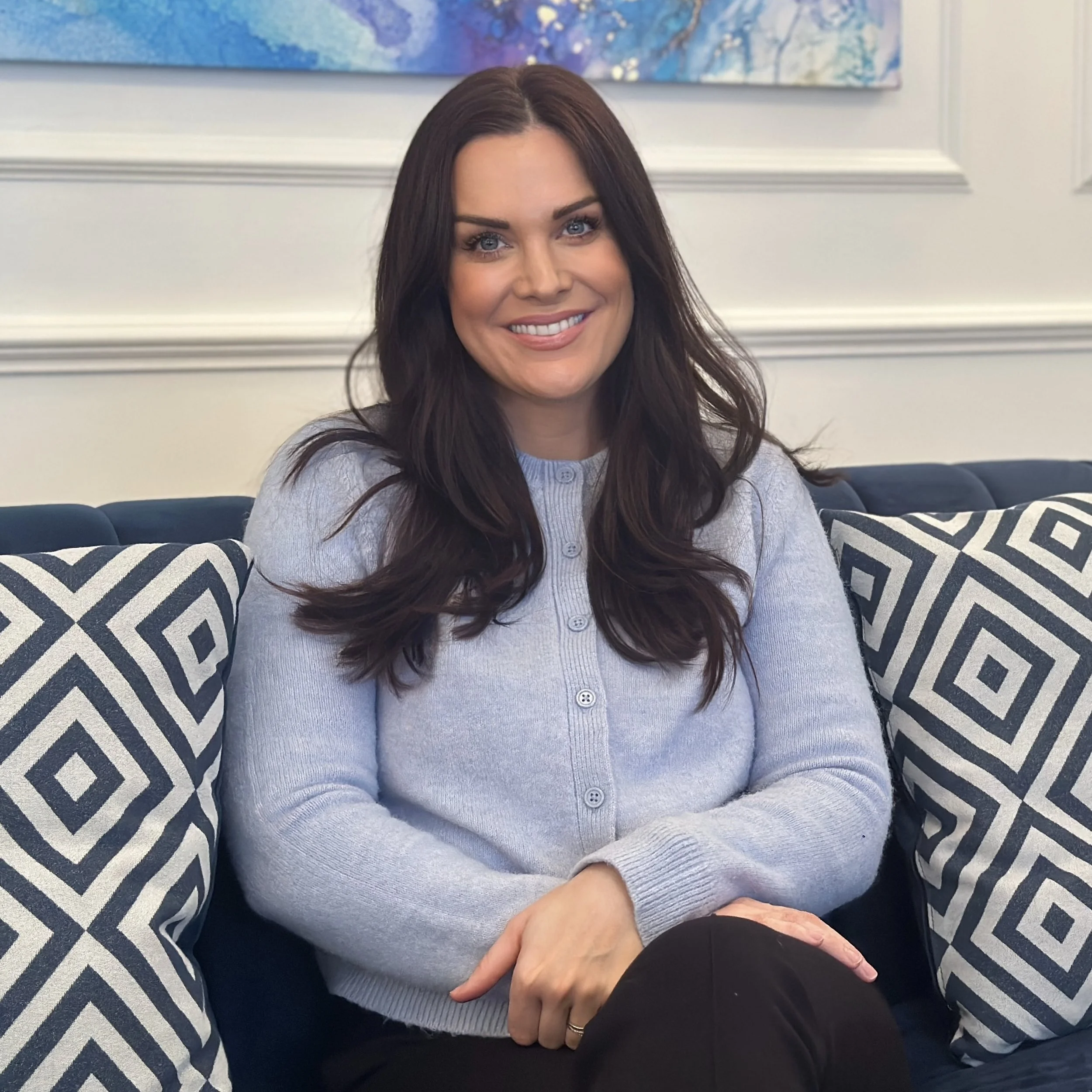 A woman with long dark hair, wearing a light blue button-up cardigan, sitting on a blue couch with patterned cushions, smiling at the camera in a room with white walls and a colorful painting in the background.