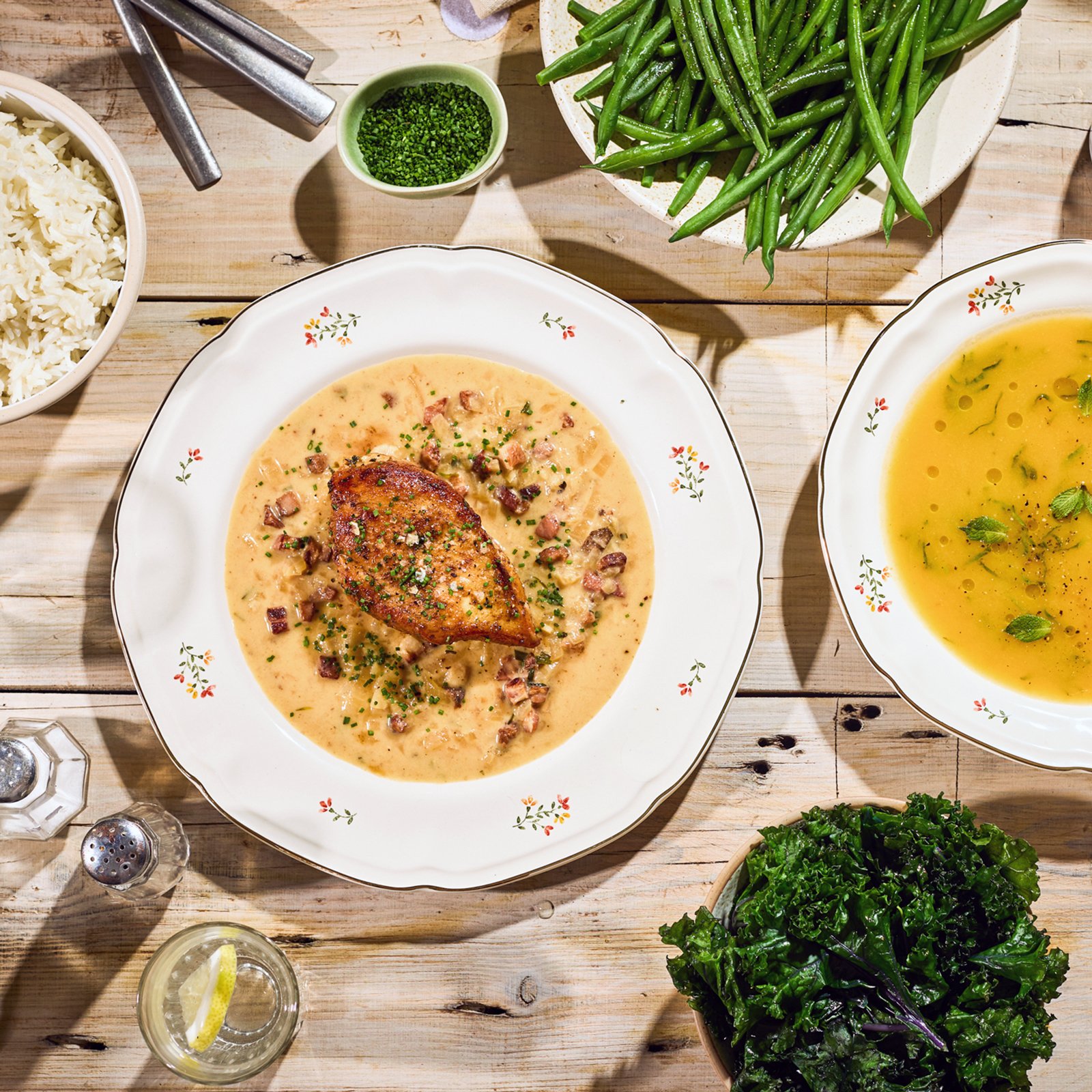 A table with a variety of dishes including a plate with chicken in creamy sauce and chopped bacon, a bowl of rice, green beans, green leafy salad, and a bowl of yellow soup garnished with herbs.