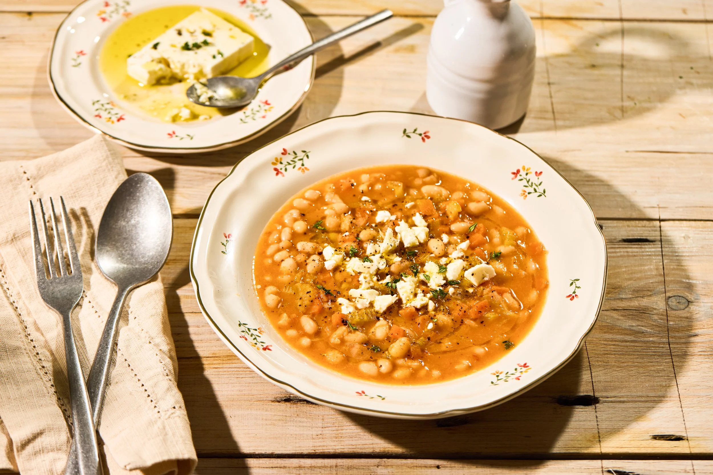 A bowl of bean soup topped with crumbled cheese and herbs on a wooden table. In the background, there is a plate with a piece of cheese in olive oil, a spoon, a salt shaker, and a fork and spoon resting on a cloth napkin.