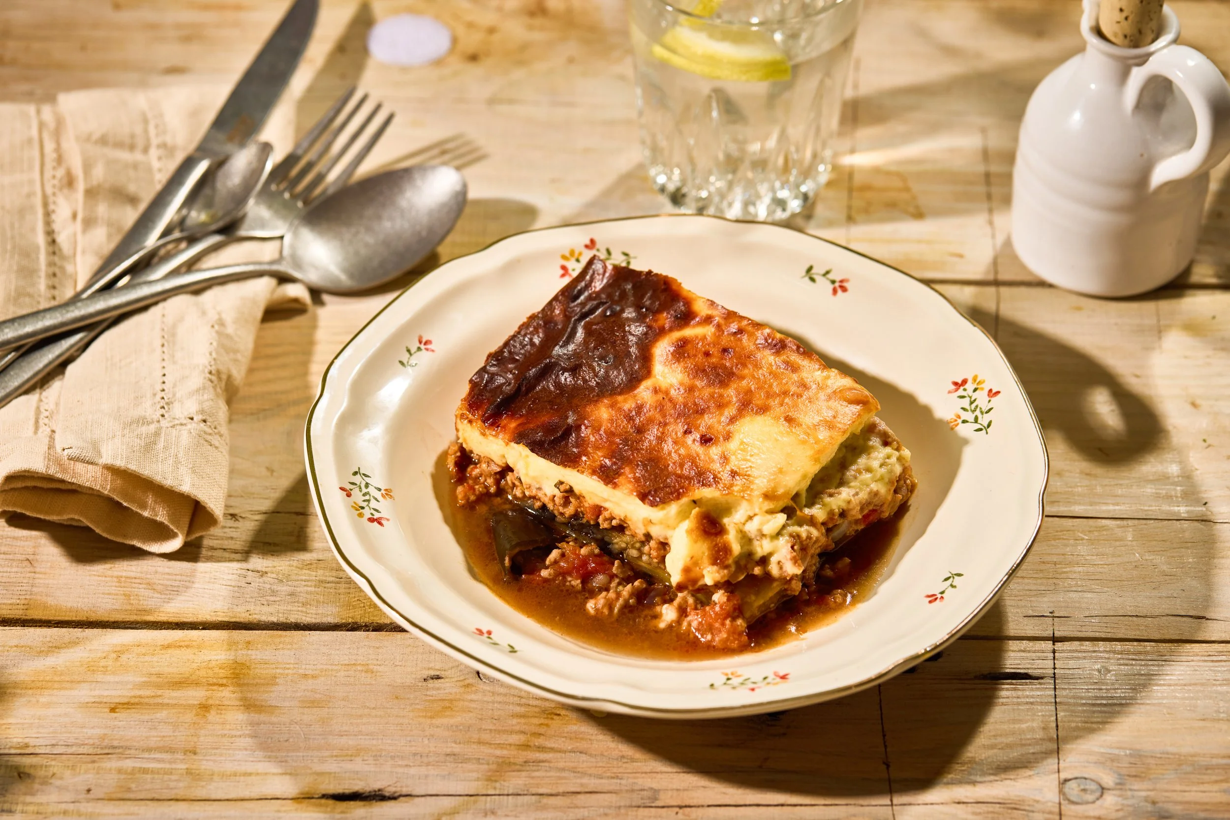 A plate of homemade lasagna on a wooden table with a glass of water with lemon slices and a small white pitcher in the background, and silverware placed on a napkin to the side.