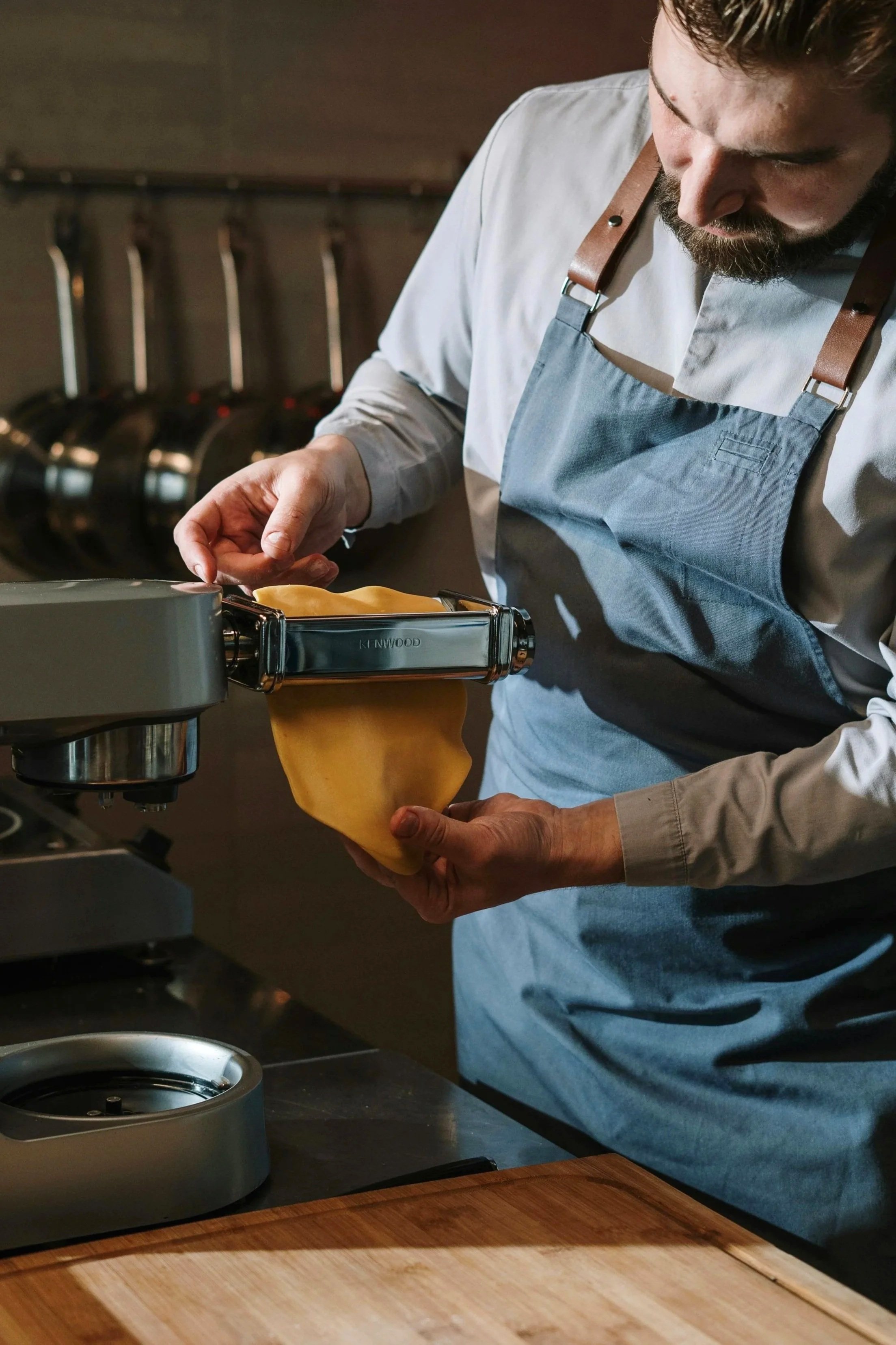 A chef in a gray apron is using an automatic pasta maker to roll out fresh dough in a kitchen.