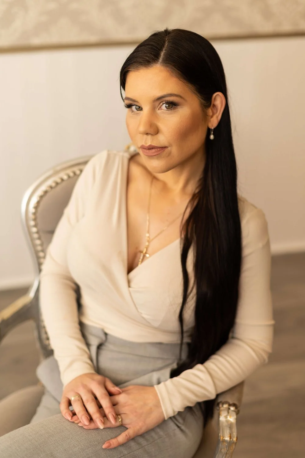 Young woman with long dark hair, dressed in a cream top and beige trousers, wearing a gold cross and chain sat in a beige chair, looking to camera