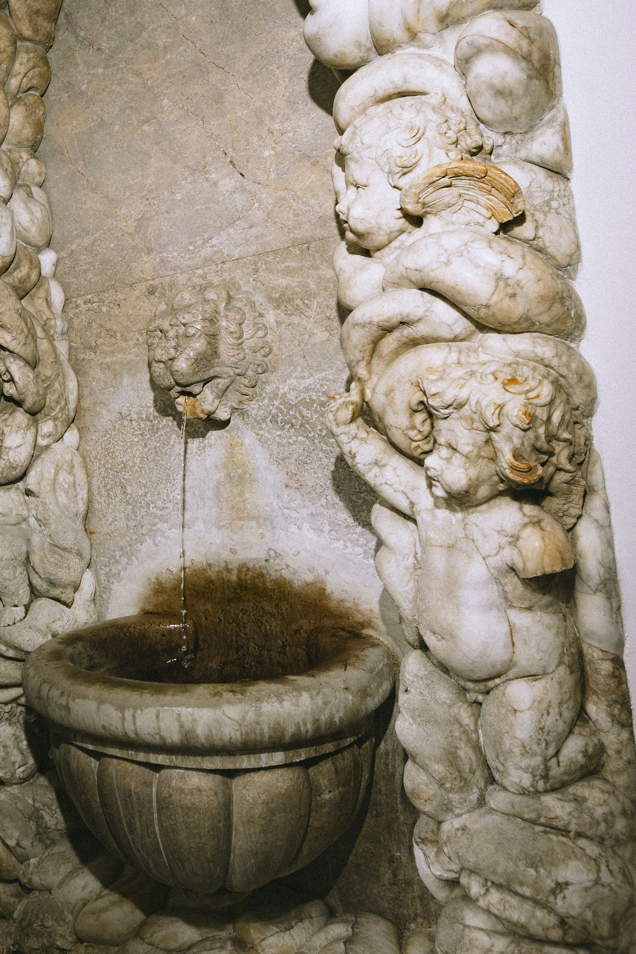 Steinfigur eines Wasserbrunnens mit Löwenkopf-Mund über einem Fass, umgeben von geschnitzten Figuren und dekorativen Elementen aus Marmor.
