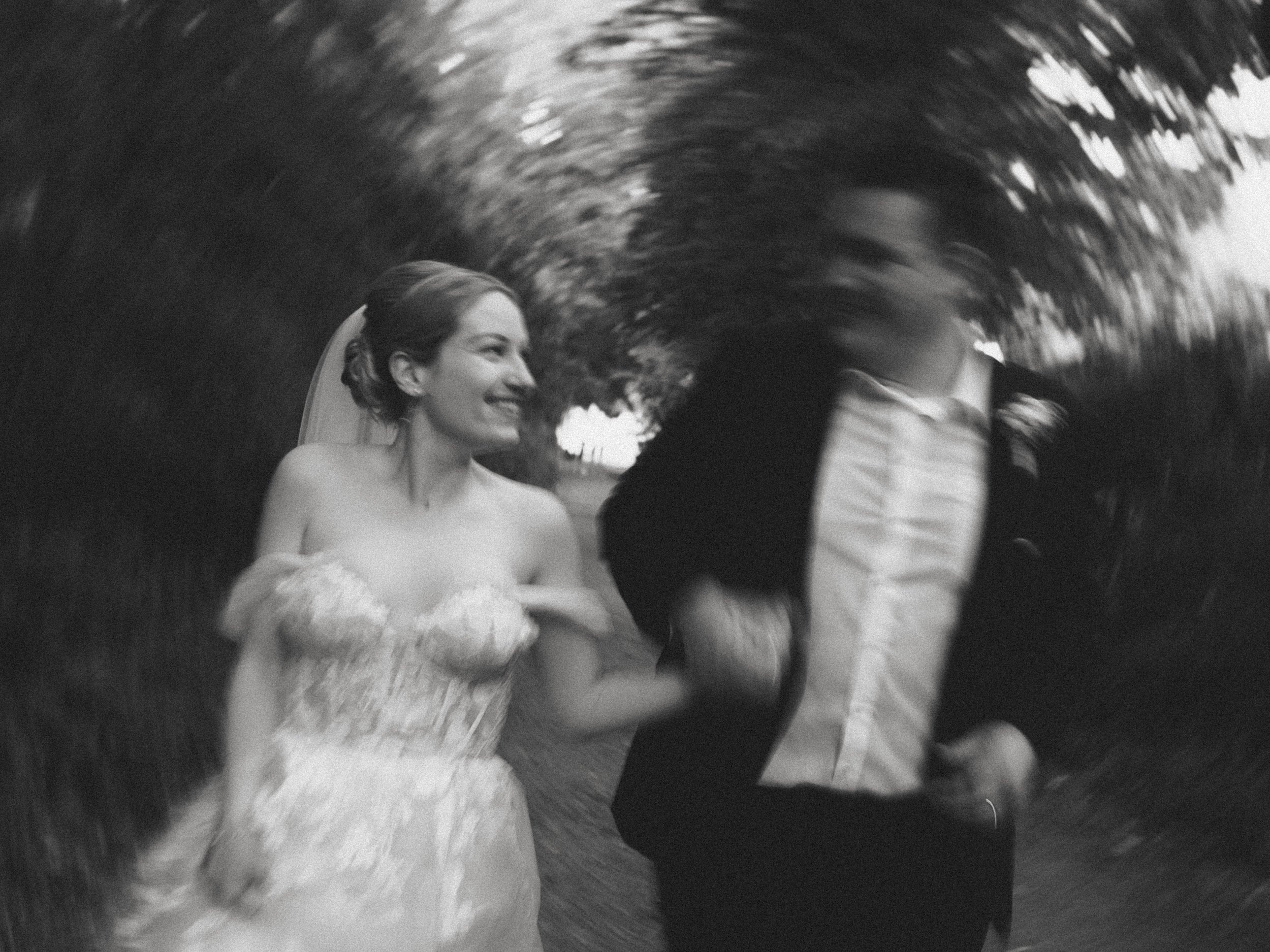 Schwarz-weiß-Foto eines lächelnden Brautpaares, bei dem die Braut ein Hochzeitskleid trägt und der Bräutigam in einem Anzug ist. Der Hintergrund zeigt eine bunte, unscharfe Naturkulisse mit Bäumen. Die Aufnahme hat einen künstlerischen, verschwommenen Effekt.