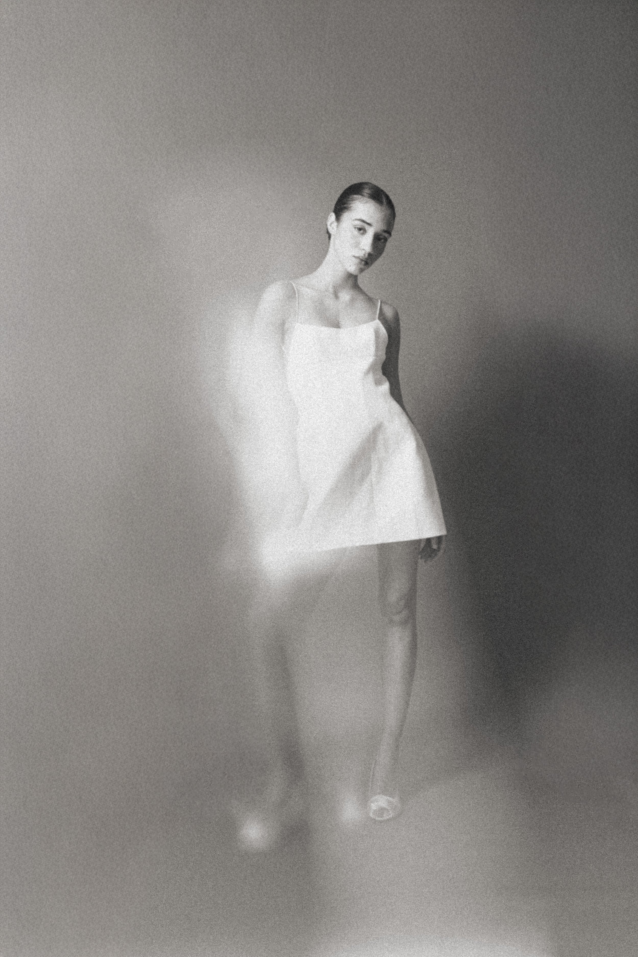 Schwarz-weiße Aufnahme einer jungen Frau in einem weißen Kleid, vor einem neutralen Hintergrund, mit einem verschwommenen, wolkenähnlichen Nebel um sie herum.