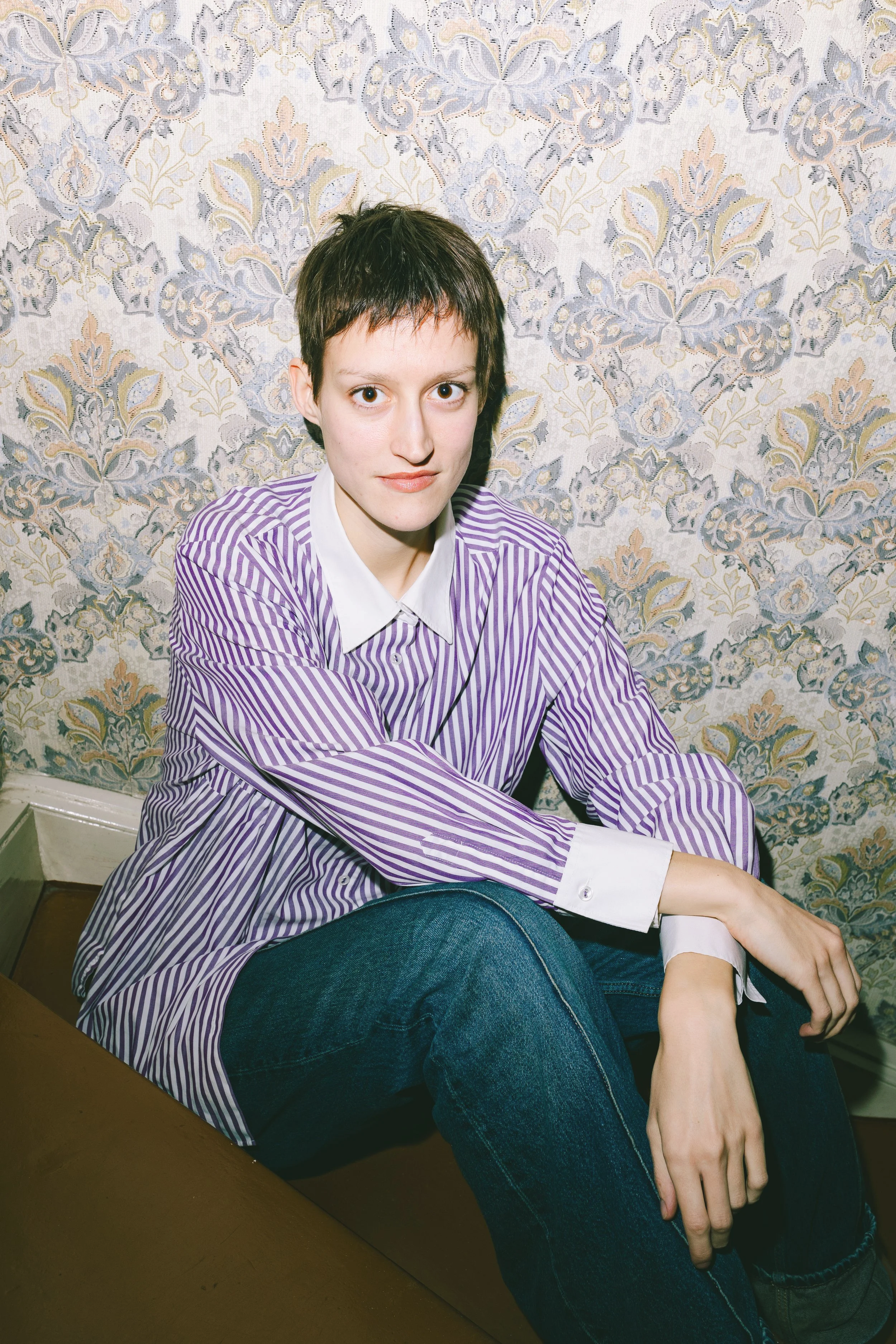 Junge Frau mit kurzer dunkler Frisur und gestreiftem Hemd sitzt im Raum vor einer gemusterten Tapete.