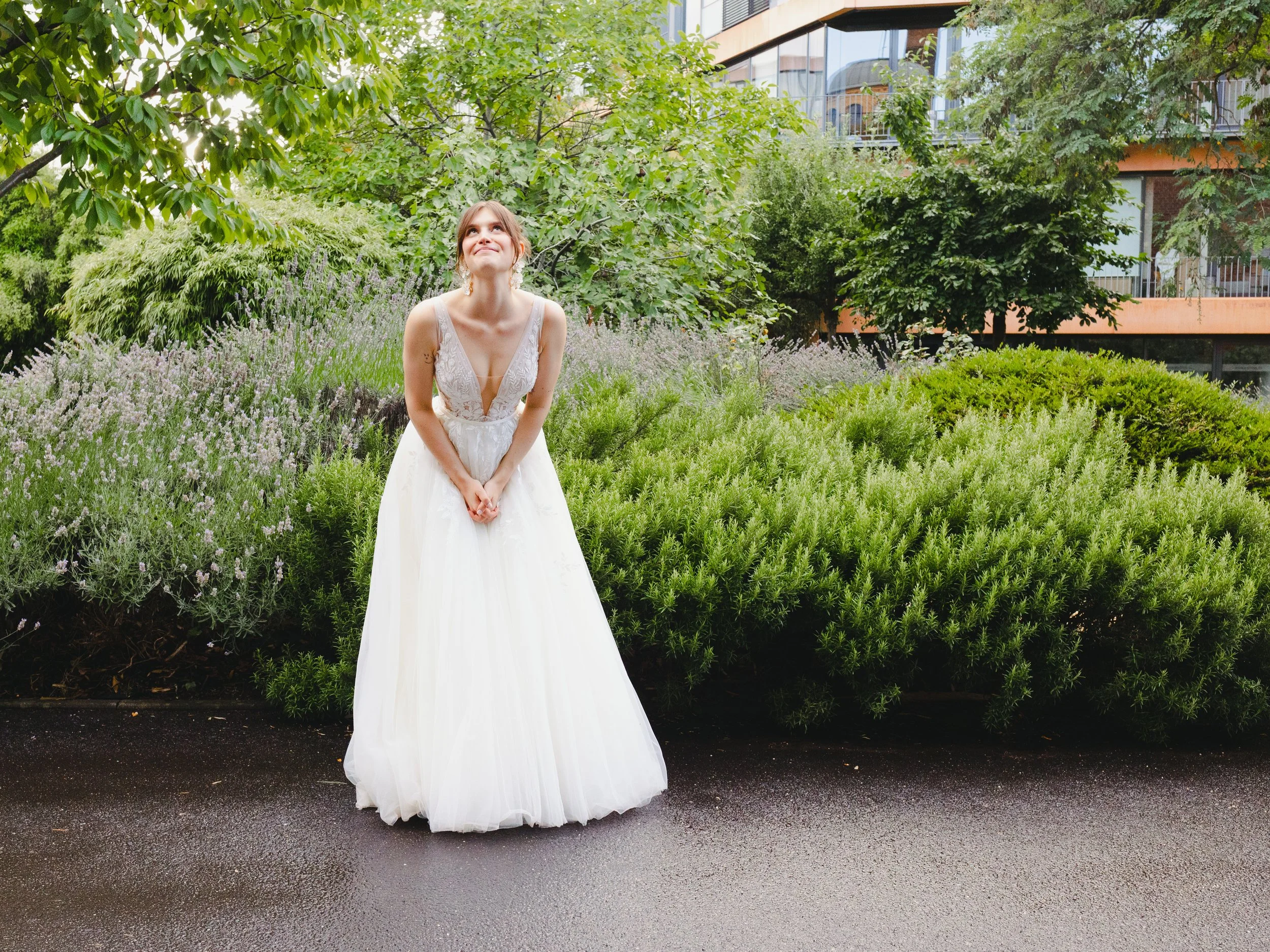 Junge Frau in einem weißen Brautkleid steht auf der Straße vor grünen Büschen und Bäumen, lächelt und blickt nach oben.