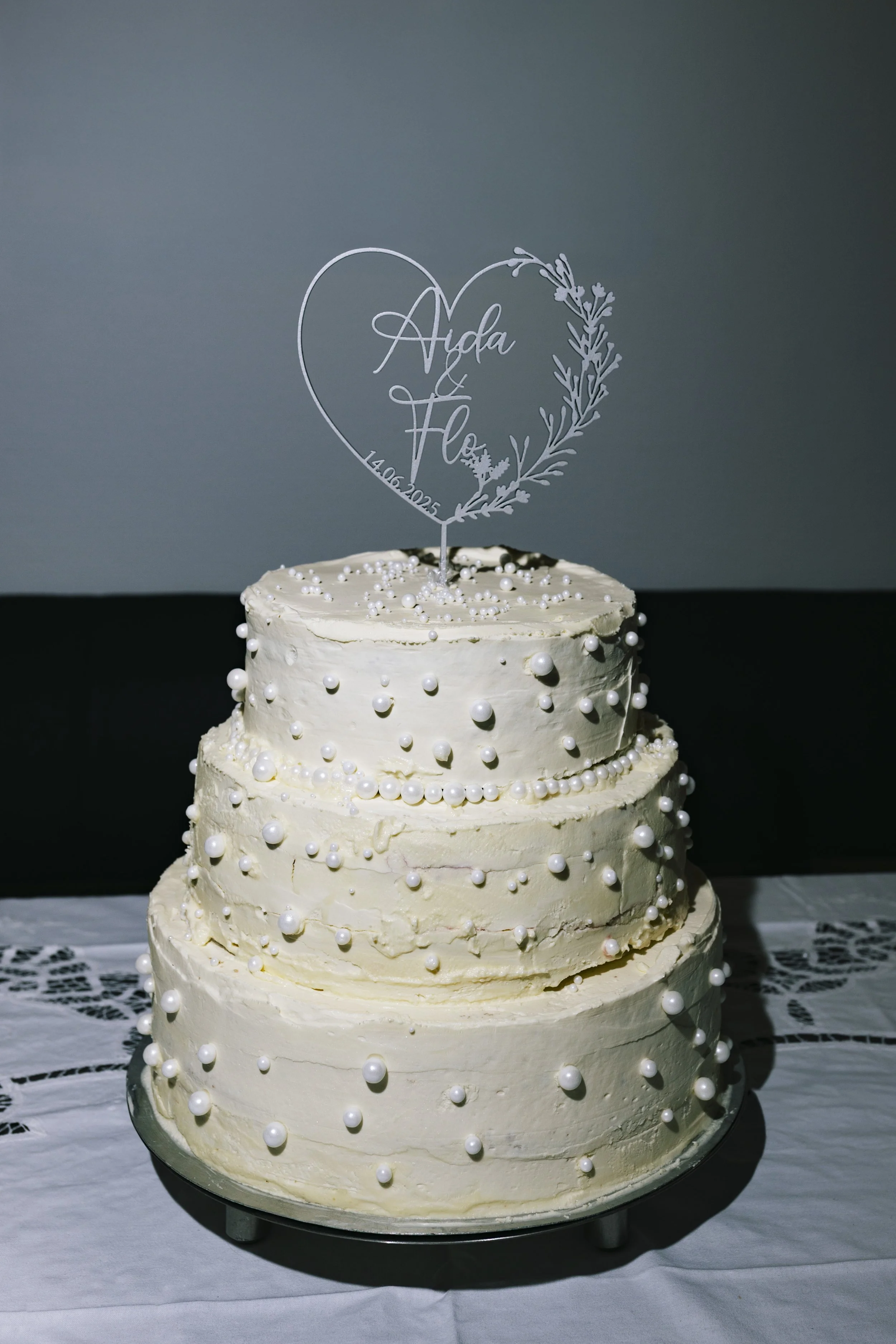 Mehrstöckige Hochzeitstorte mit weißen Perlen und einem Herz-Topper mit den Namen 'Ada' und 'Flo' sowie dem Datum '14.06.2023'.