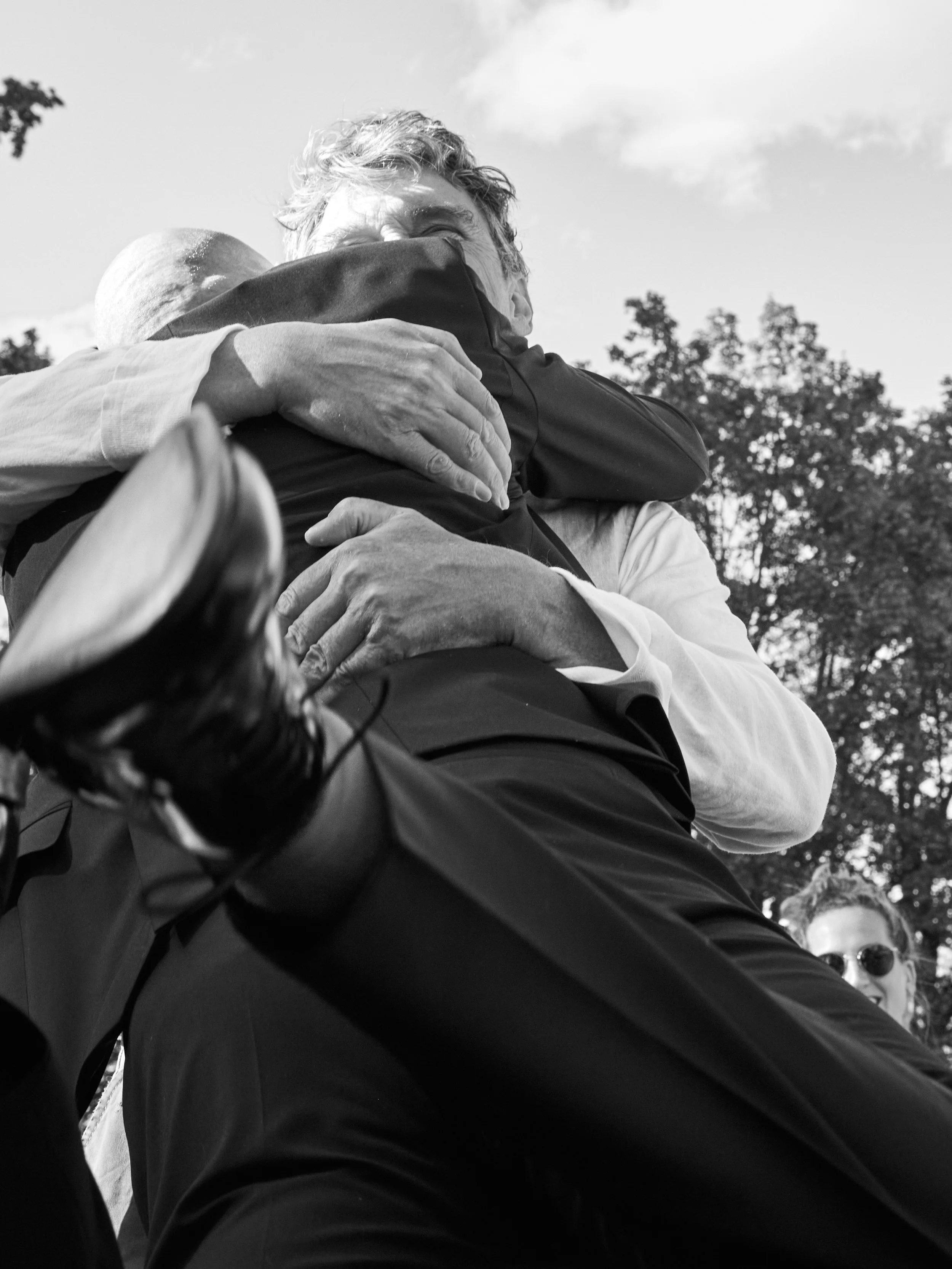 Zwei Menschen umarmen sich leidenschaftlich, eines in einem weißen Hemd, das andere in dunkler Kleidung, im Freien mit Bäumen im Hintergrund, schwarz-weiß Foto.