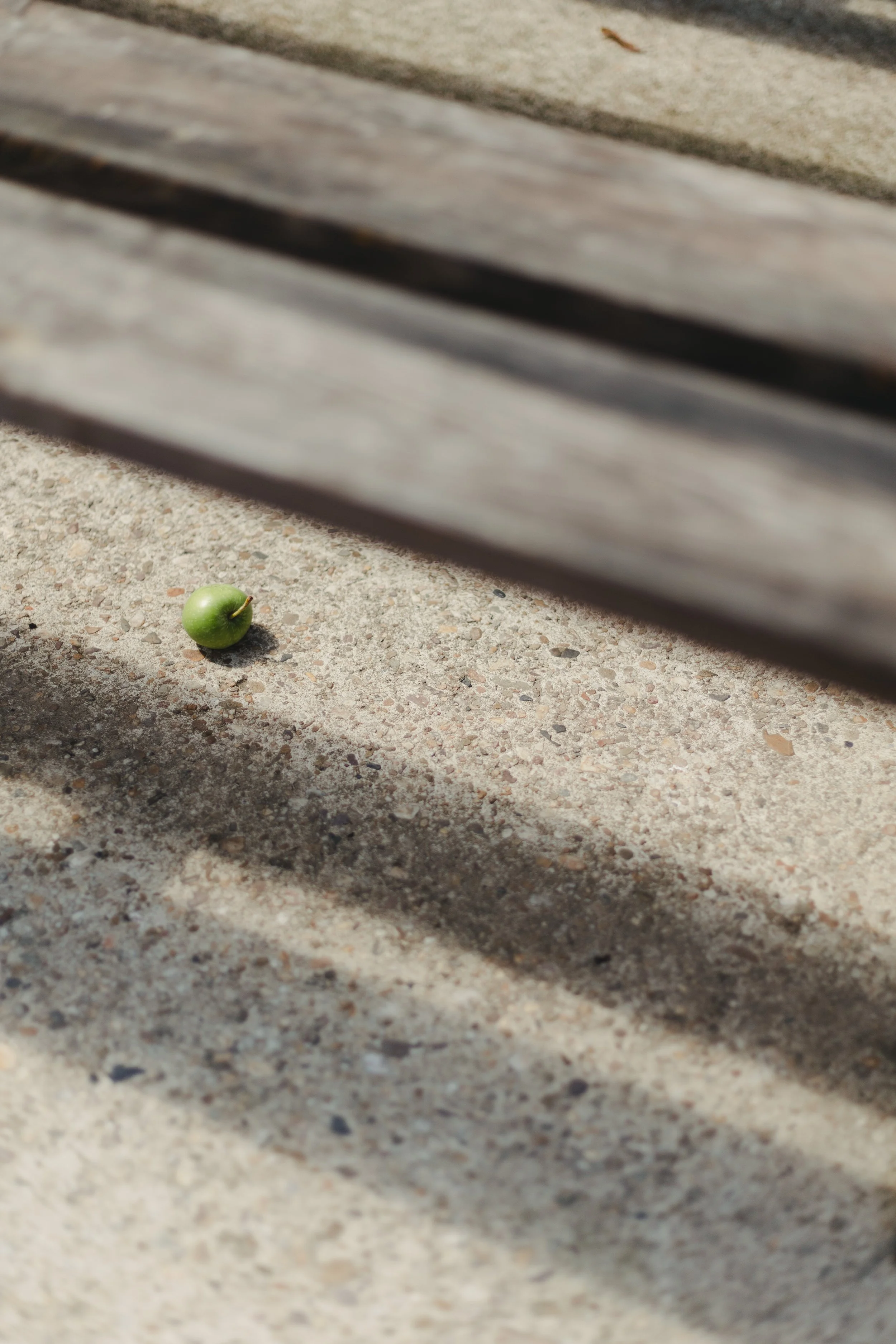 Kleiner grüner Apfel auf Betonboden neben einer Holzbank, mit Schatten und Sonnenlicht.