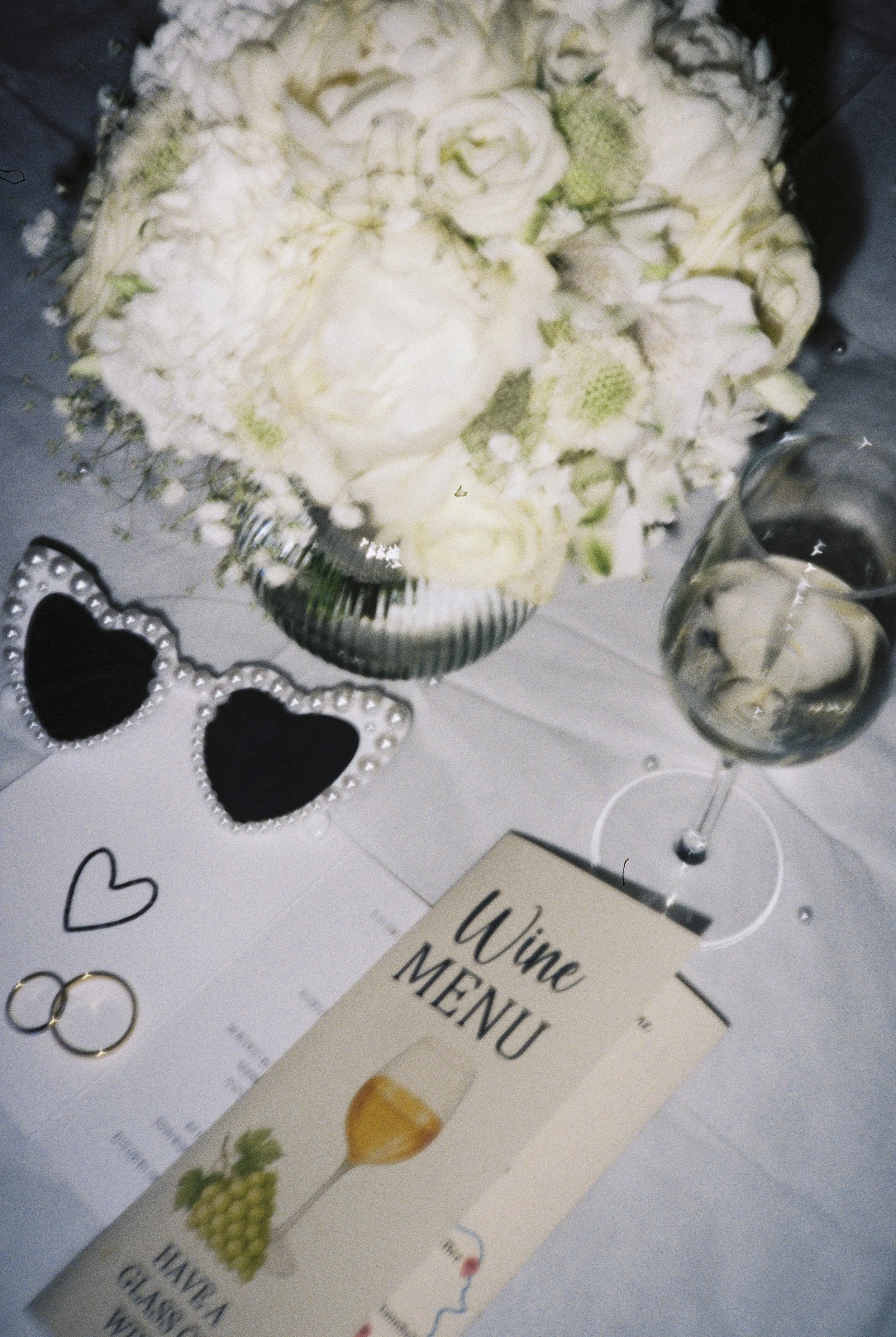 Blumenstrauß mit weißen Rosen und anderen weißen Blumen, zwei Weingläser mit Wein, eine Hochzeitsdekoration mit zwei schwarzen Herzen mit Perlenrändern, Goldringen, eine Wein-Menu-Karte, ein weißer Tisch.