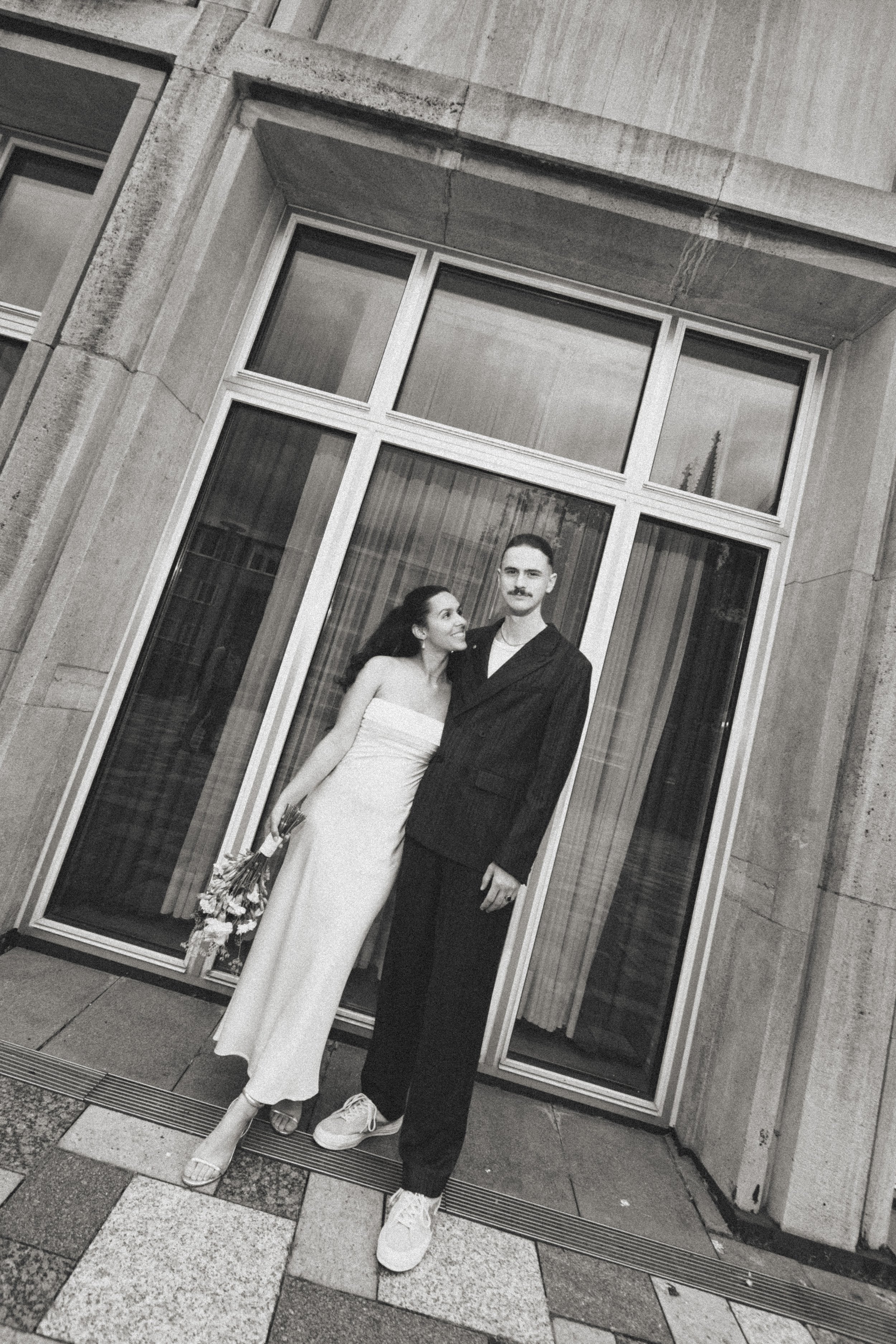 Ein Schwarz-Weiß-Foto eines Paares, das auf einer Straße vor einem Gebäude mit großen Fenstern steht. Die Frau trägt ein langes, straffes Kleid und hält einen Blumenstrauß, während der Mann einen dunklen Anzug trägt. Beide blicken liebevoll zueinande