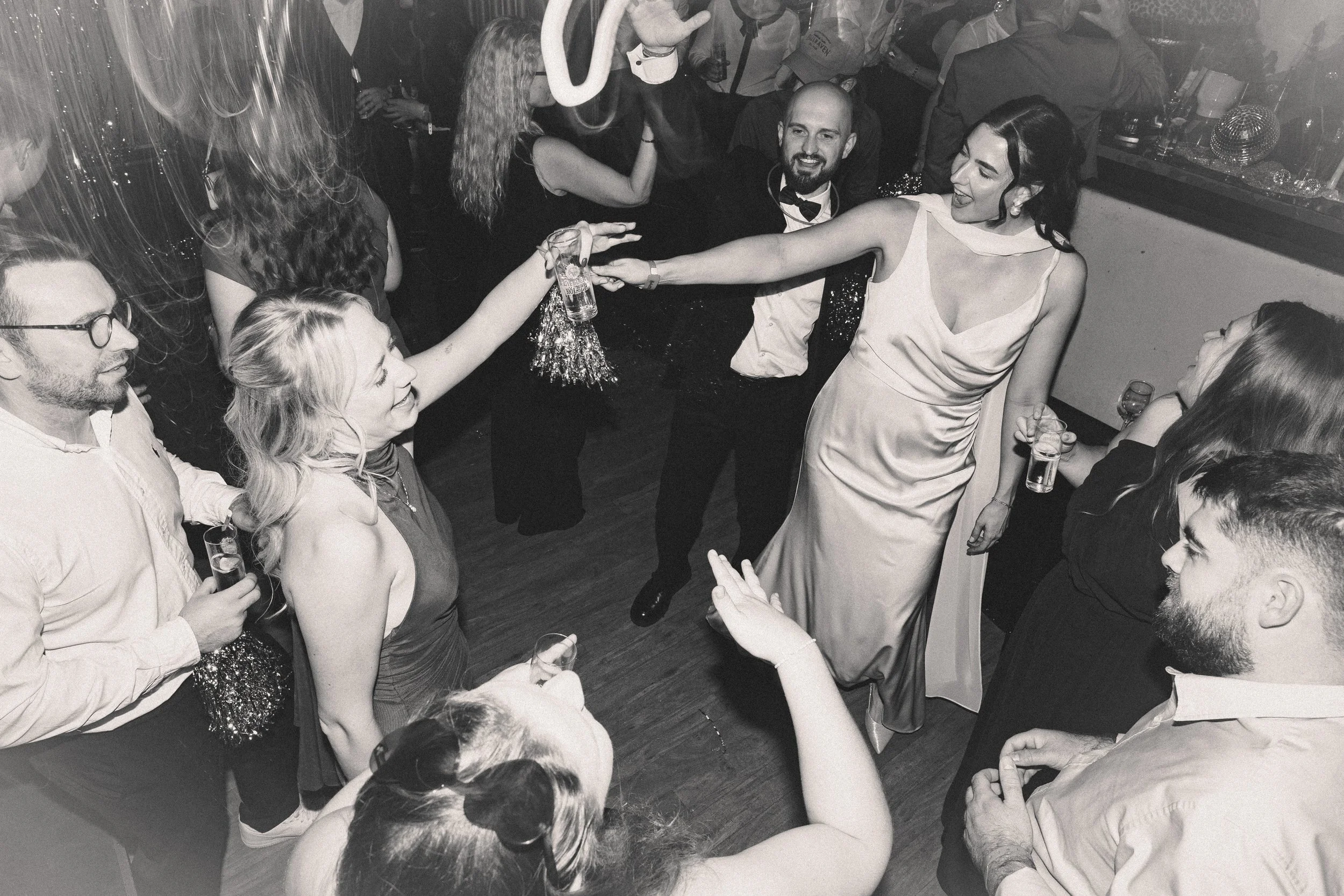 Menschen feiern auf einer Party, einige halten Getränke in der Hand, lachen und tanzen, im Hintergrund weitere Gäste und Dekorationen.