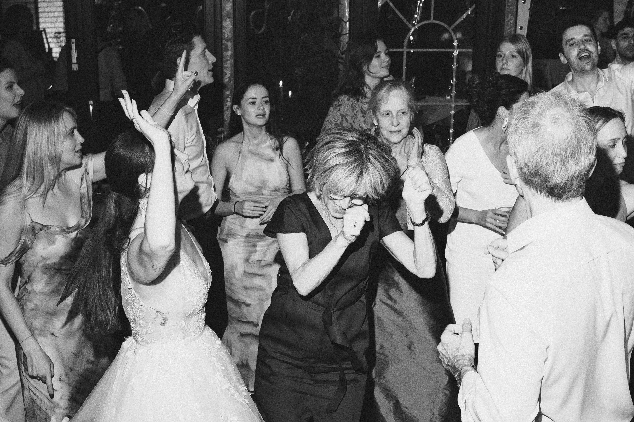 Menschen feiern auf einer Party, einige lachen und tanzen, im Vordergrund eine Frau mit Brille und schwarzen Kleid, umgeben von anderen Leuten in festlicher Kleidung, in schwarz-weiß