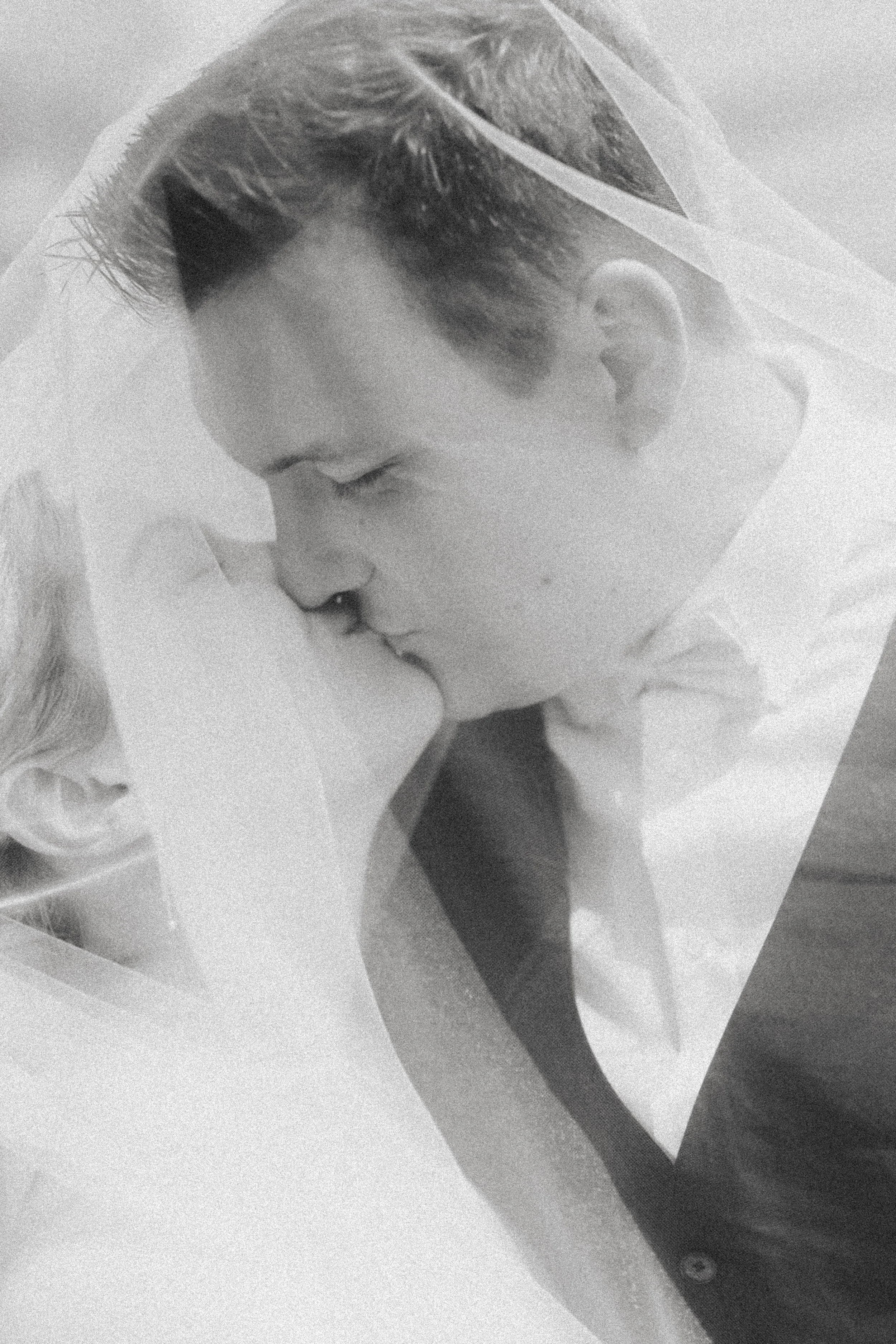 Ein Mann und eine Frau küssen sich liebevoll, beide tragen Hochzeitskleidung, die Szene ist in Schwarz-Weiß.