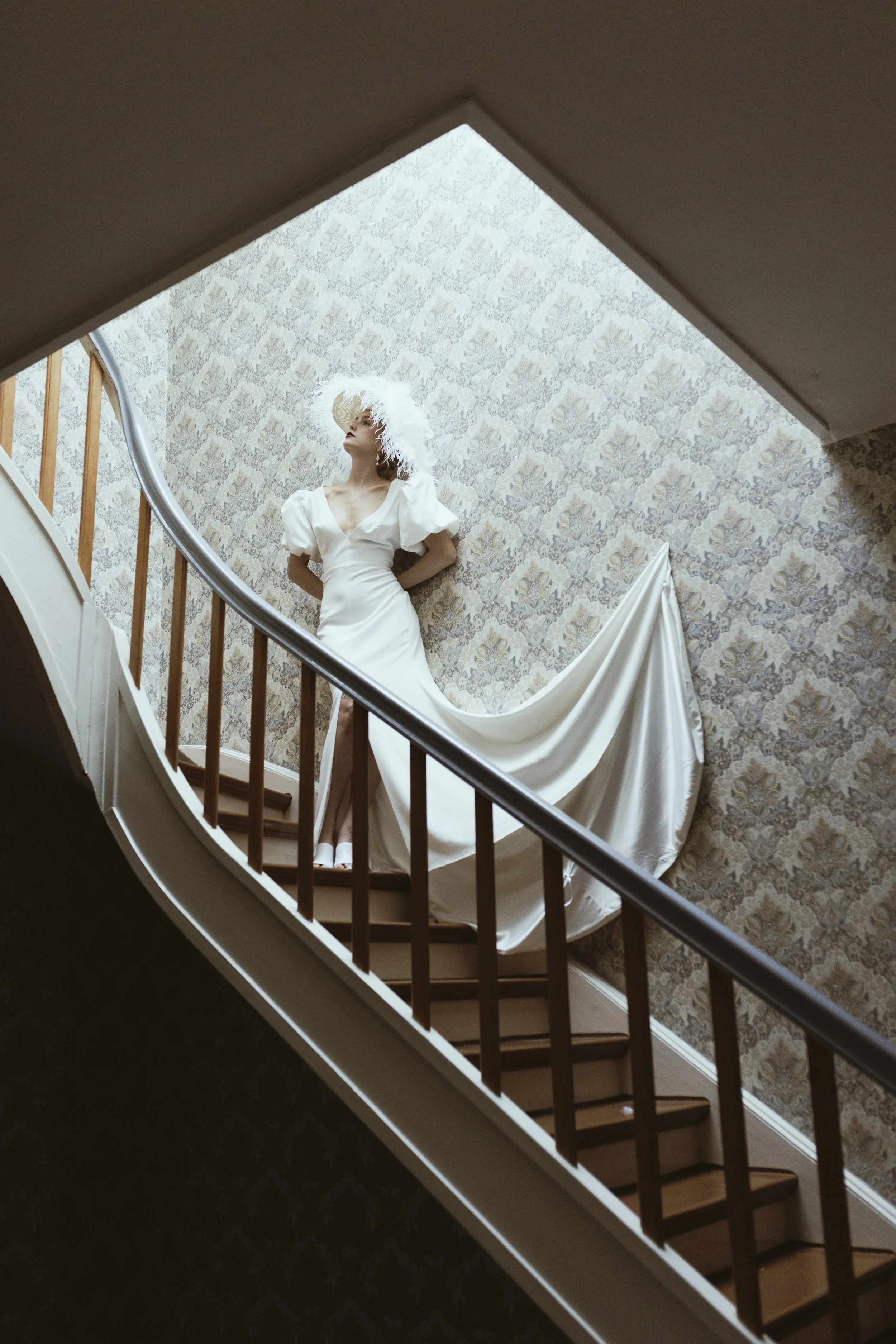 Frau in weißem Hochzeitskleid mit Kopfputz auf heikler Treppe fotografiert, im Hintergrund alte Tapete.