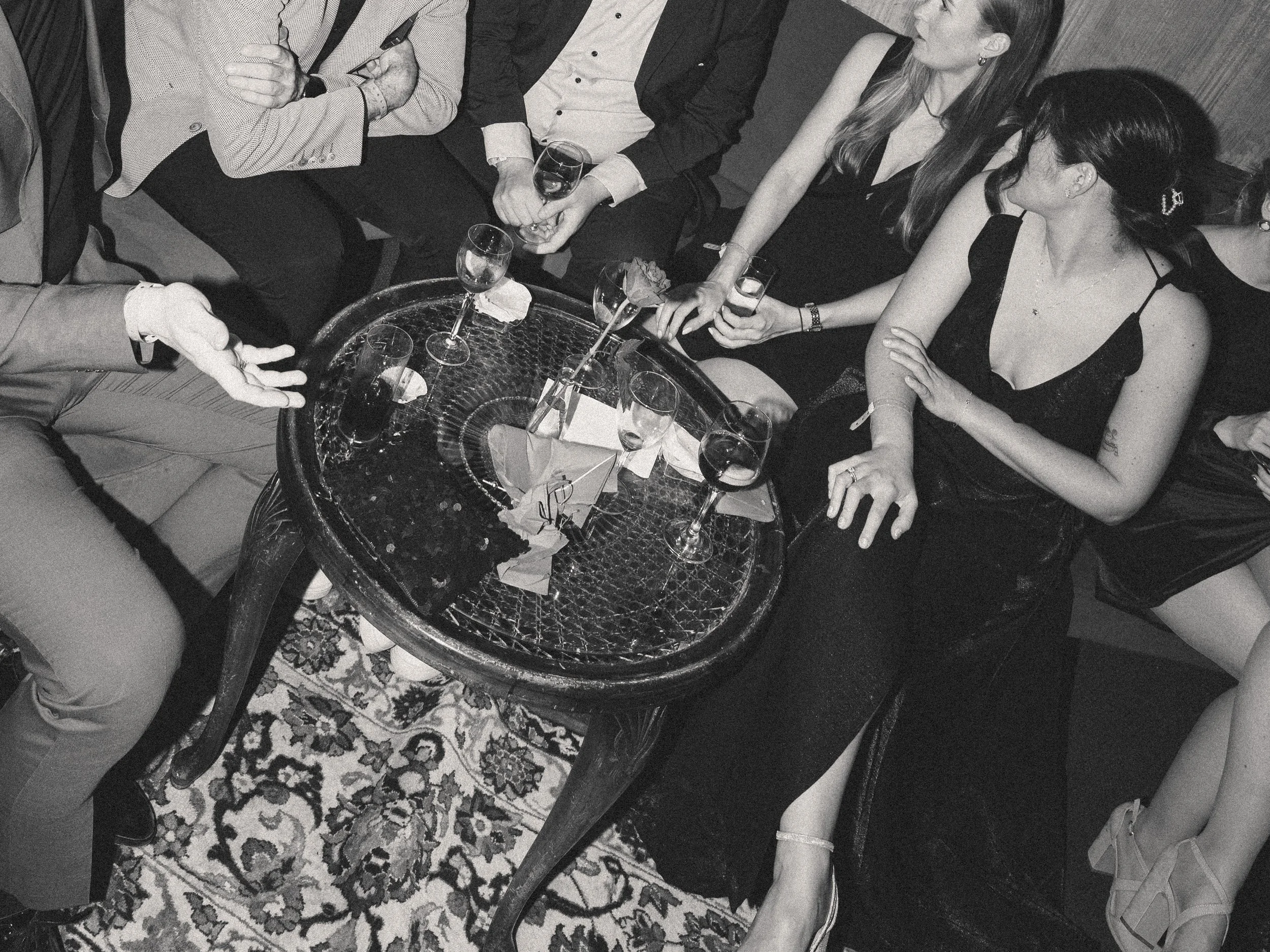 Menschen sitzen um einen runden Tisch mit leeren und halb vollen Weingläsern, auf einem Sofa und einem Stuhl, bei einer gesellschaftlichen Zusammenkunft.