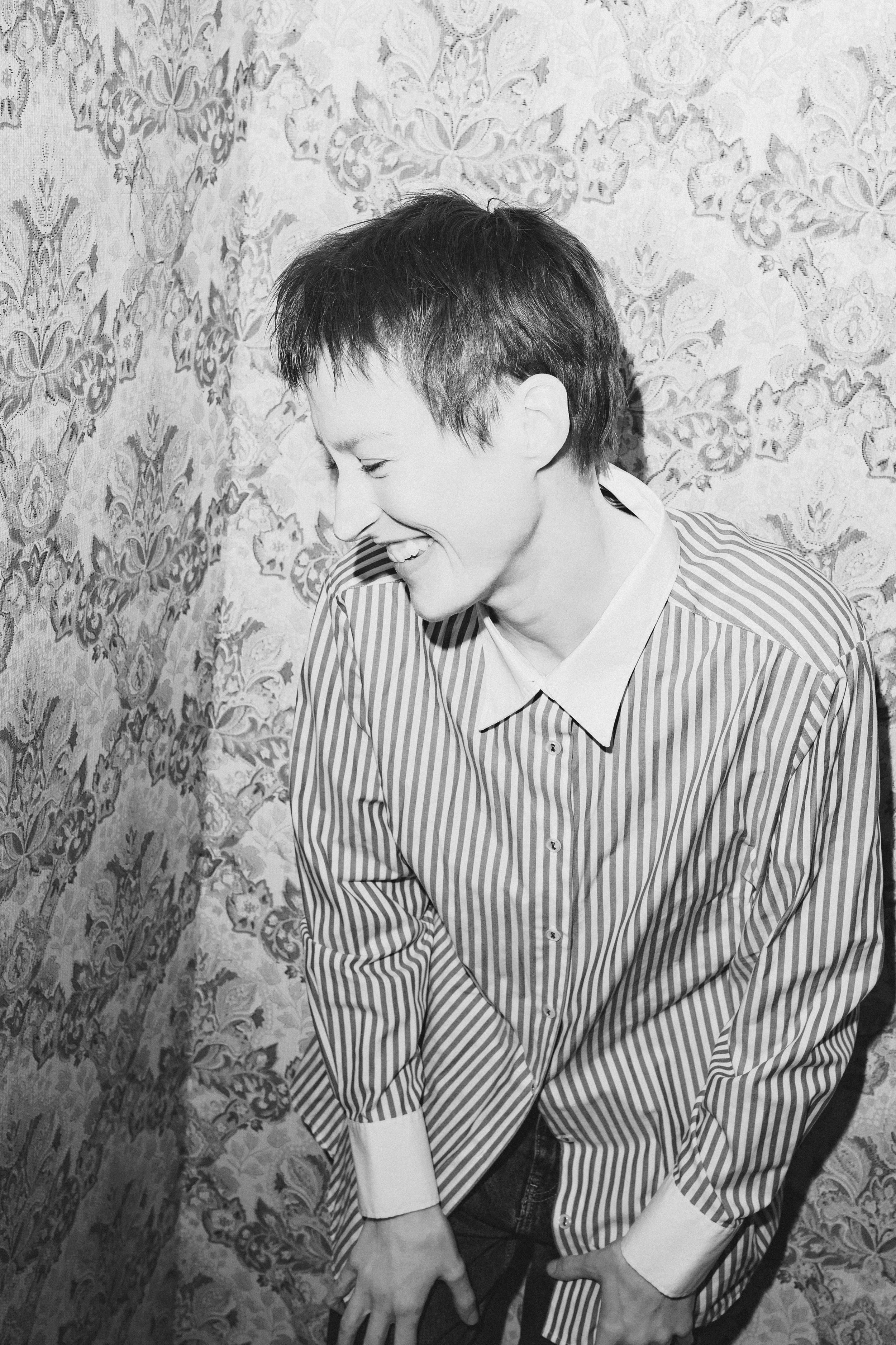 Junge Frau mit kurzem Haar trägt ein gestreiftes Hemd, lacht und schaut nach unten, vor einer gemusterten Wand.