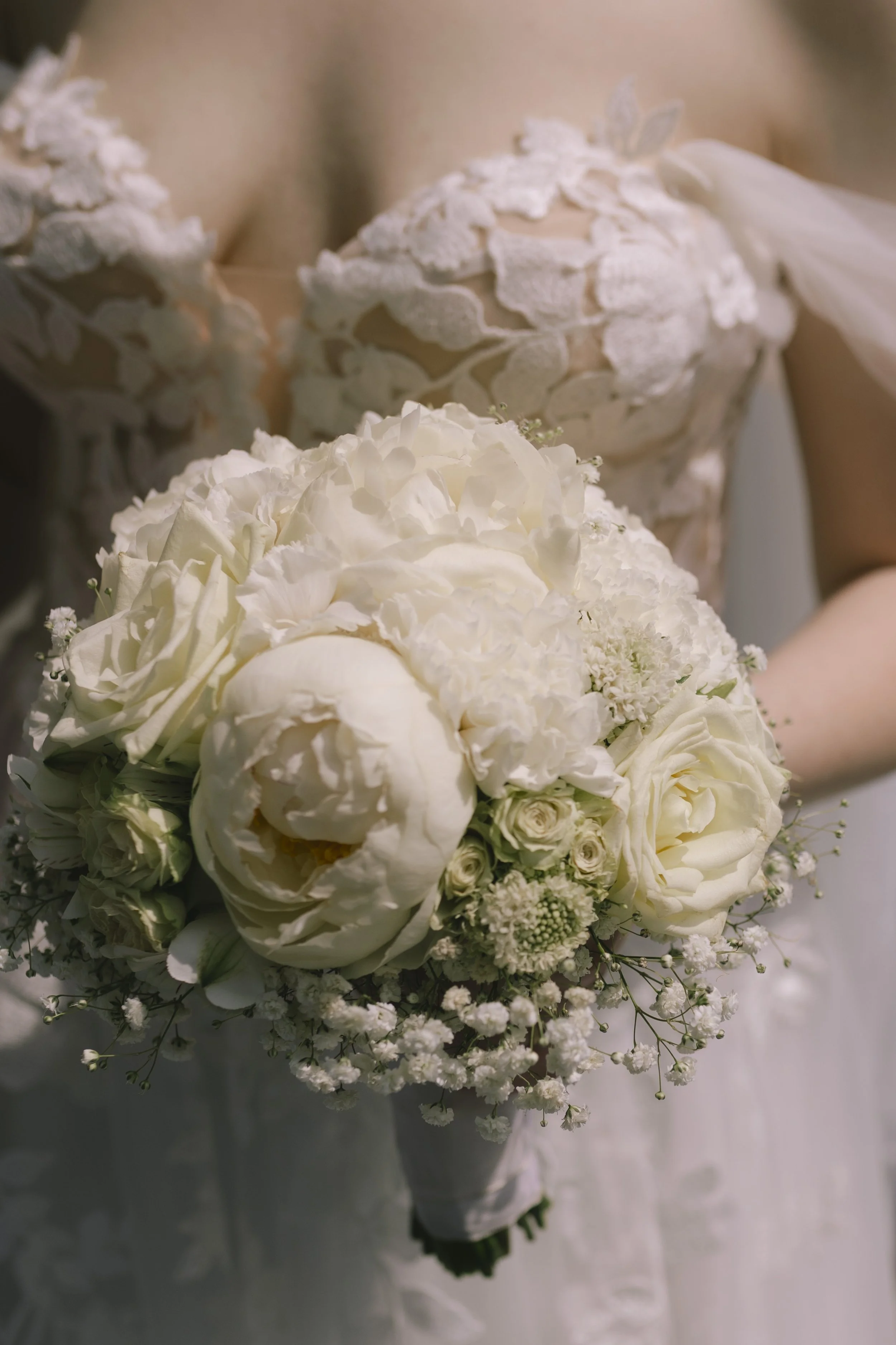 Eine Braut hält einen weißen Hochzeitsstrauß mit Rosen, Pfingstrosen und kleinen weißen Blumen, vor einem Brautkleid mit Spitze.