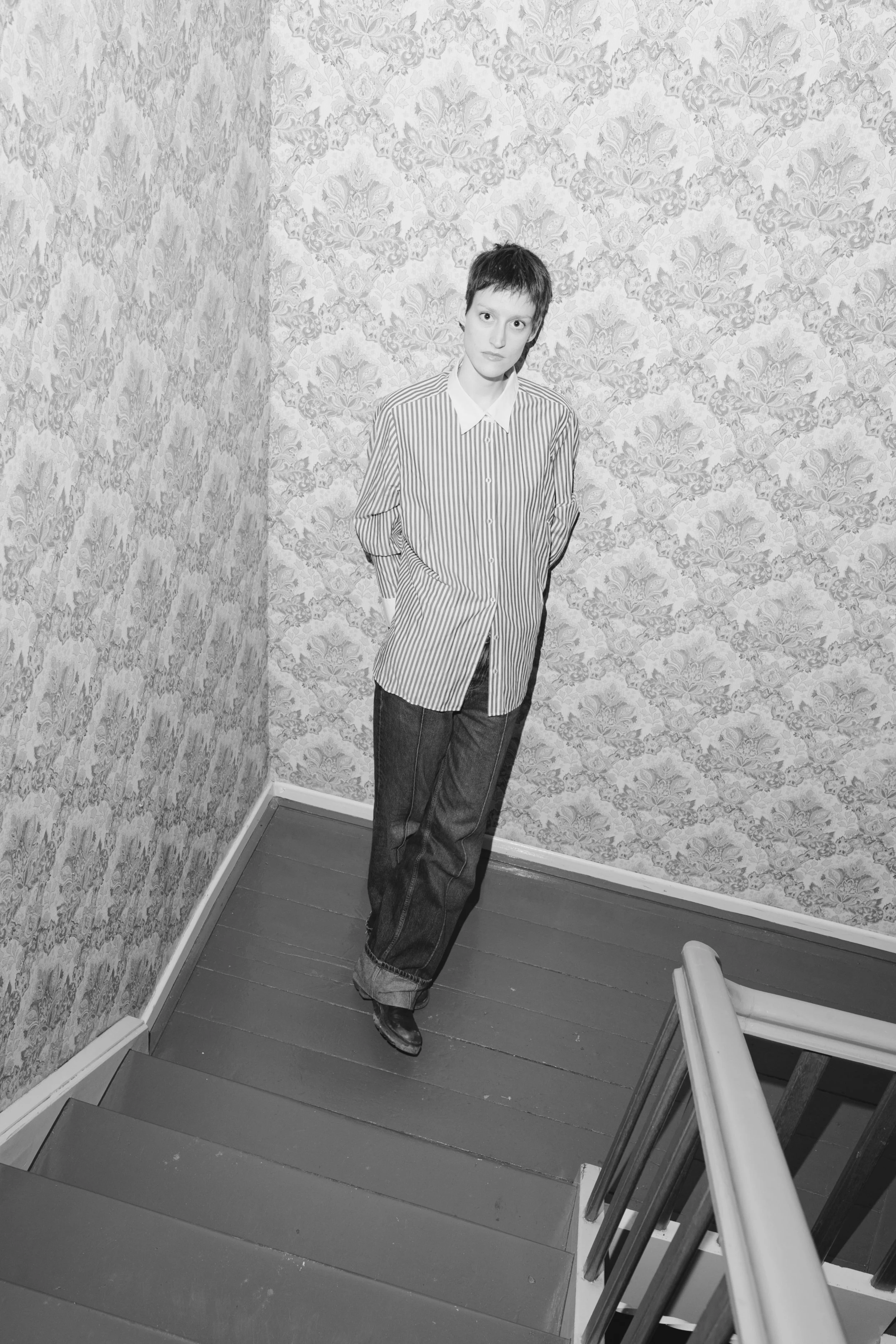 Junge Frau mit kurzen Haaren in gestreiftem Hemd und Jeans, steht auf einer Treppe in einem Raum mit Blumenmuster an den Wänden.