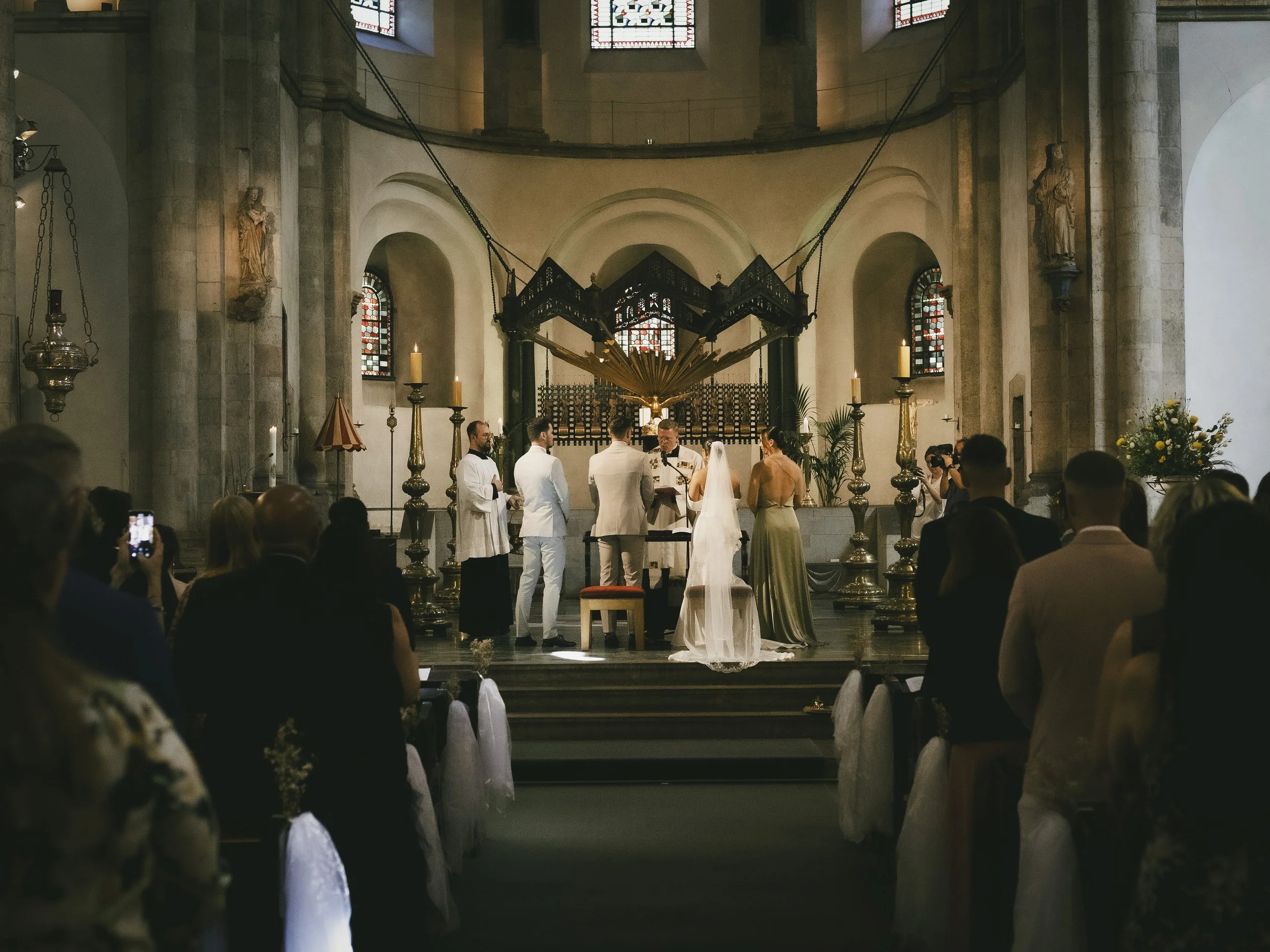 Eine Hochzeitszeremonie in einer Kirche mit Braut, Bräutigam, Trauzeugen und Pfarrer, im Vordergrund sitzen Gäste, die die Zeremonie beobachten.