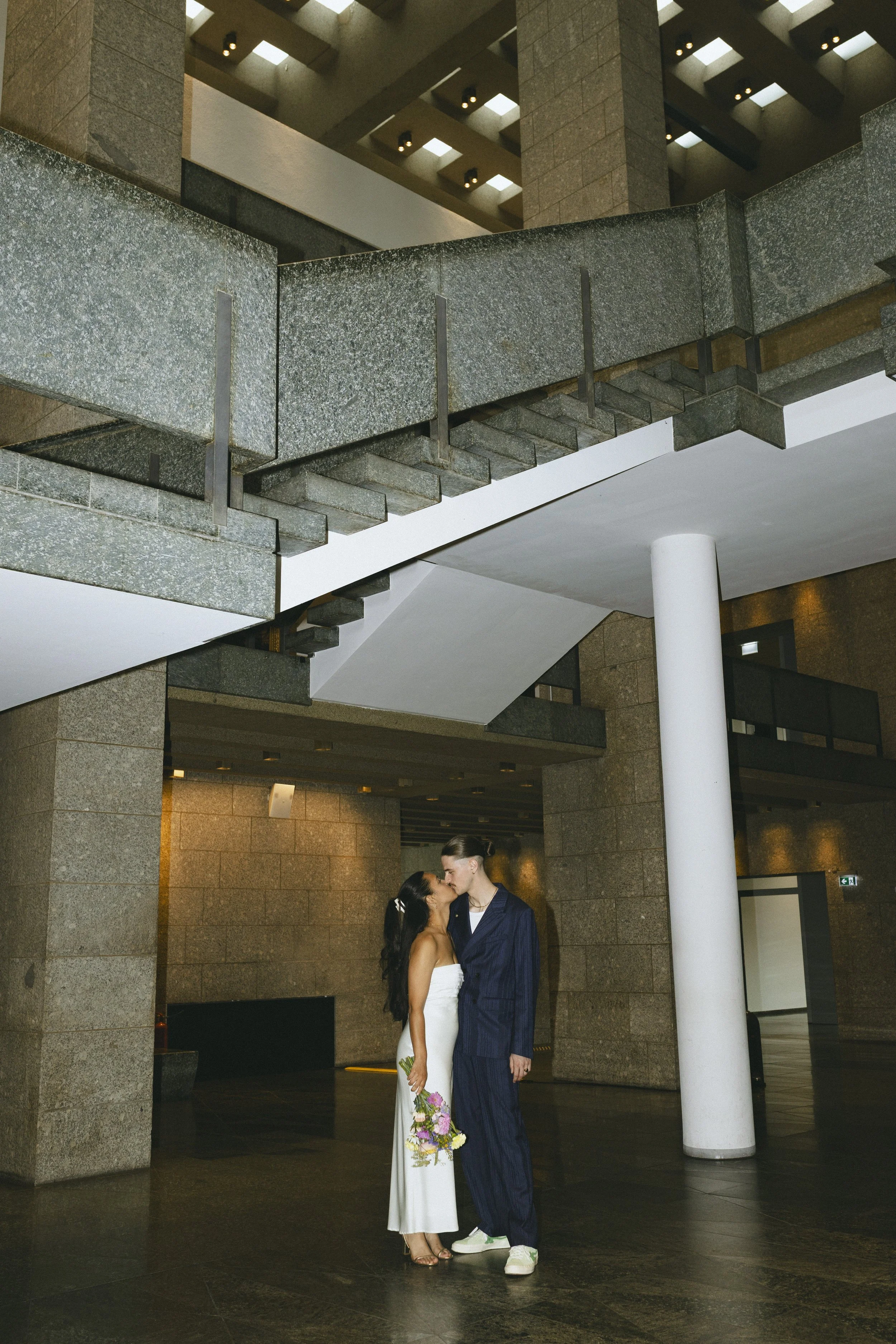 Ein Paar bei einer Hochzeit in einem modernen, mehrstöckigen Gebäude, das sich küsst. Die Frau trägt ein weißes Kleid und hält einen Blumenstrauß, der Mann trägt einen dunklen Anzug.