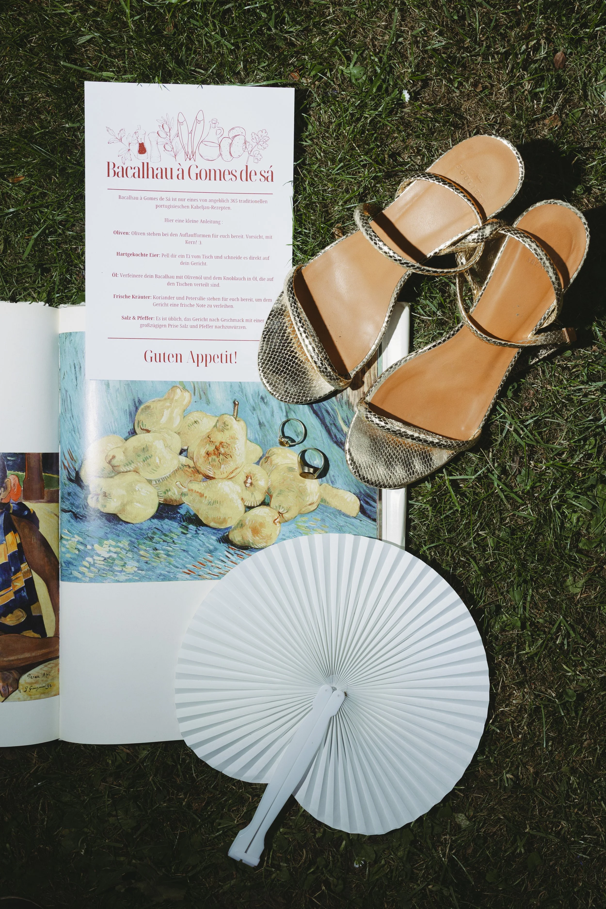 Ein offener Reiseführer mit einem Rezept für Bacalhau à Gomes de Sá, eine Karte, ein Paar Schuhe mit Absatz, zwei Ringe und ein Fächer liegen auf Gras.