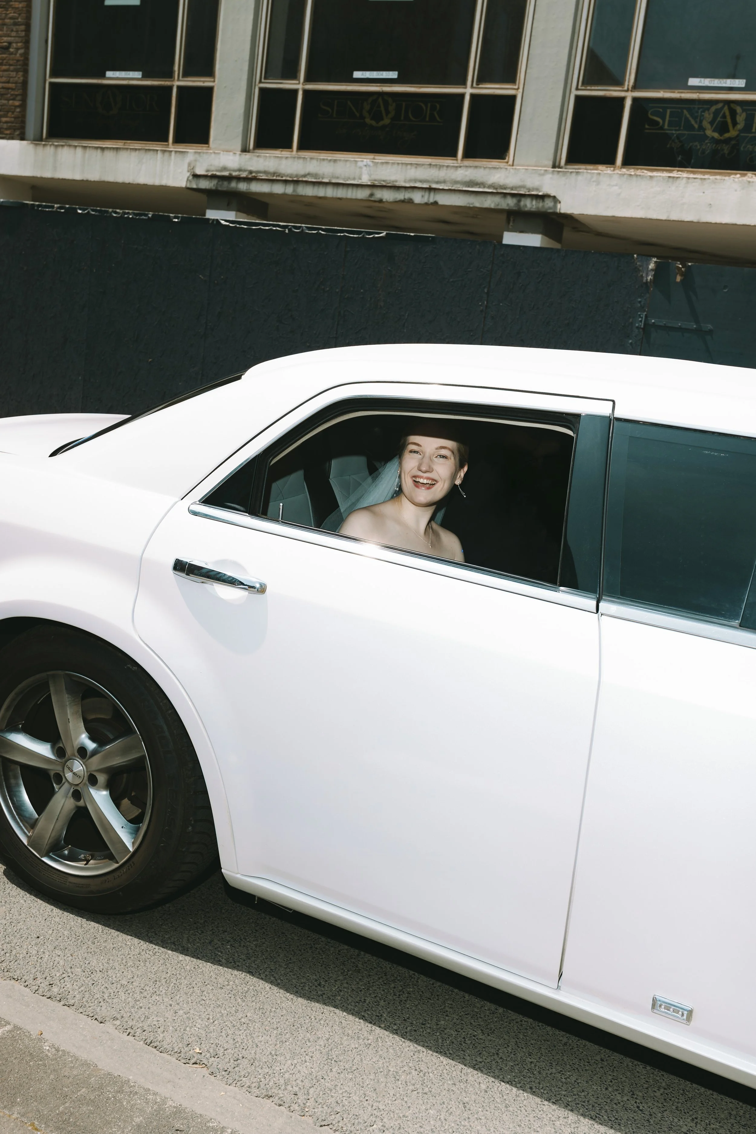 Junge Frau im weißen Hochzeitskleid sitzt in einem weißen Auto und lacht.