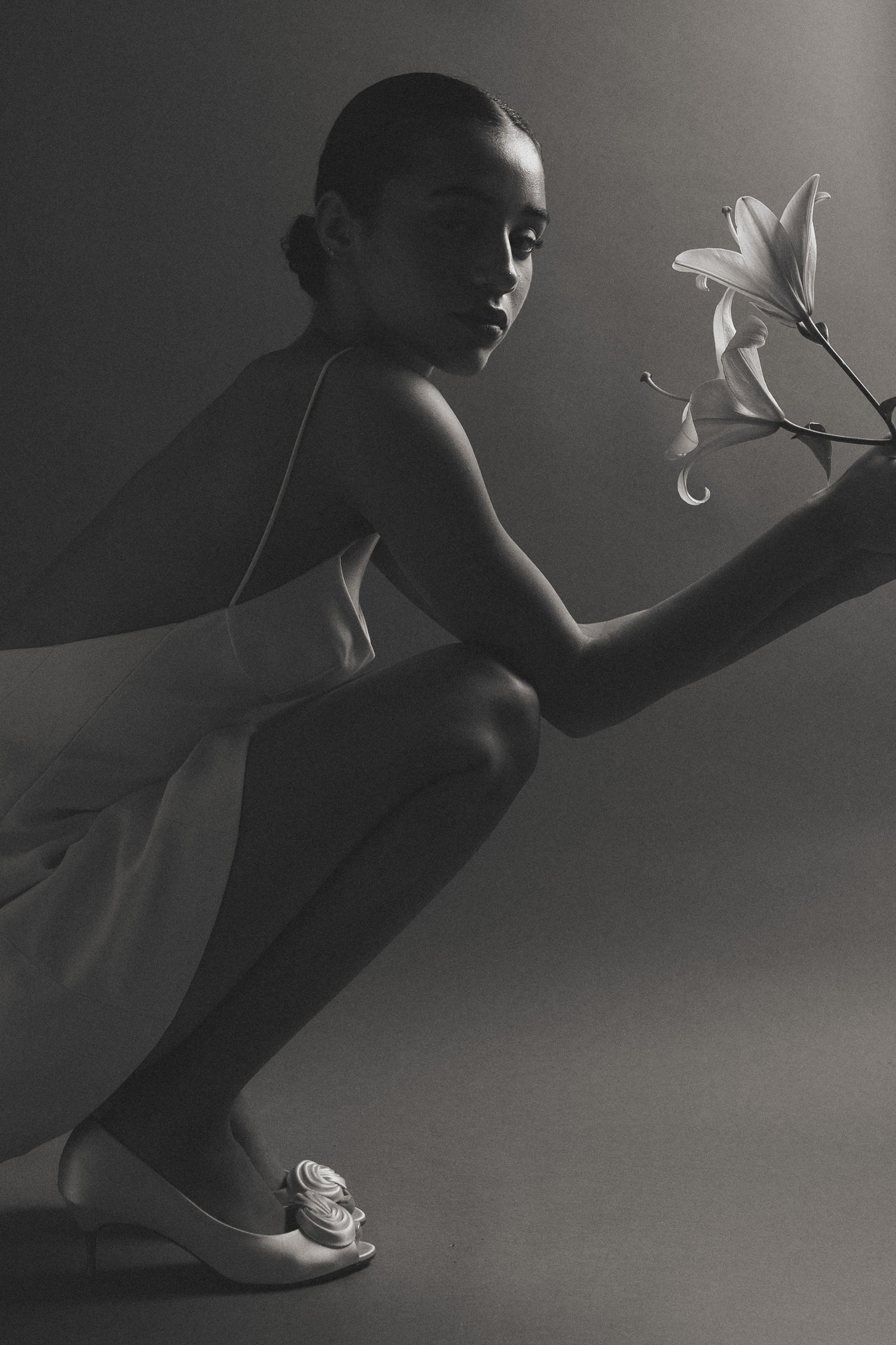 Schwarz-weiß-Fotografie einer Frau im knienden Hocke mit weißen Schuhen, einem Kleid und einer Lilie in der Hand.