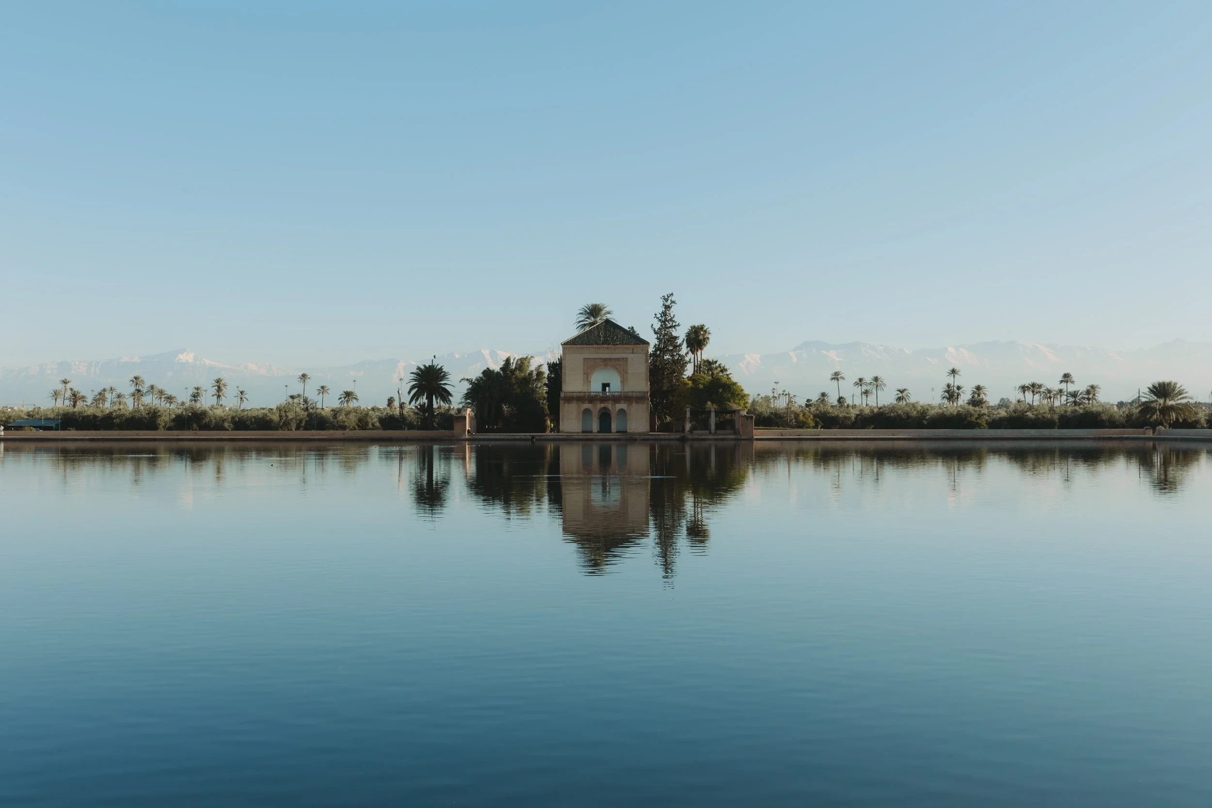Ein ruhiger See mit Reflexion eines kleinen, historischen Gebäudes, umgeben von Palmen, mit Bergen im Hintergrund bei klarem Himmel.