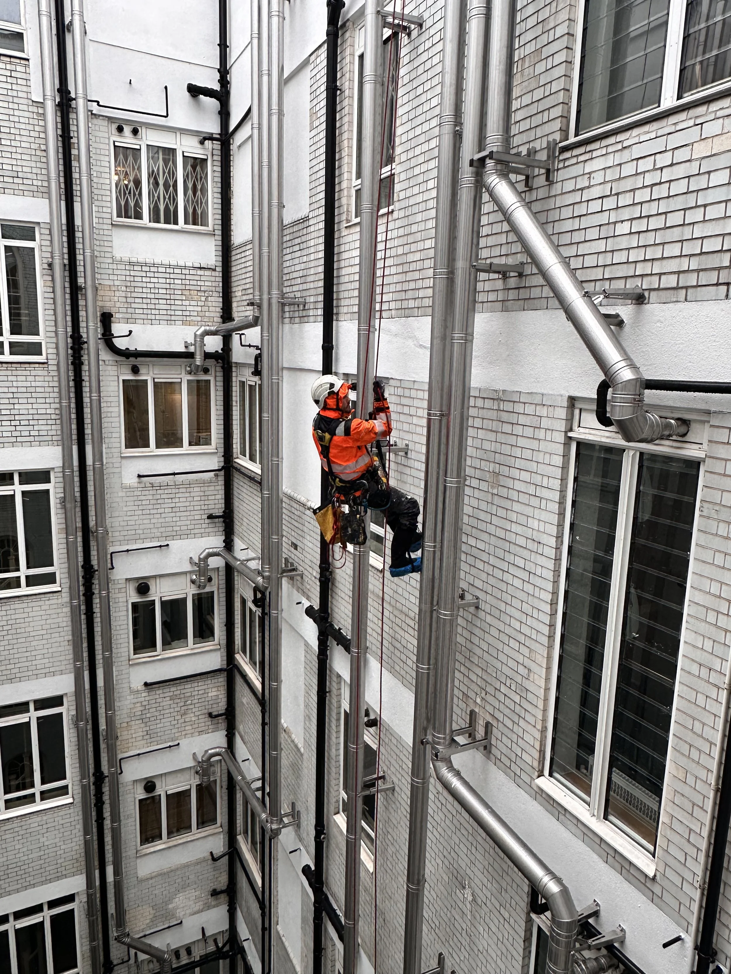 Worker in safety gear cleaning windows on a multi-story brick building using an industrial window washing system with metal brackets and support cables.
