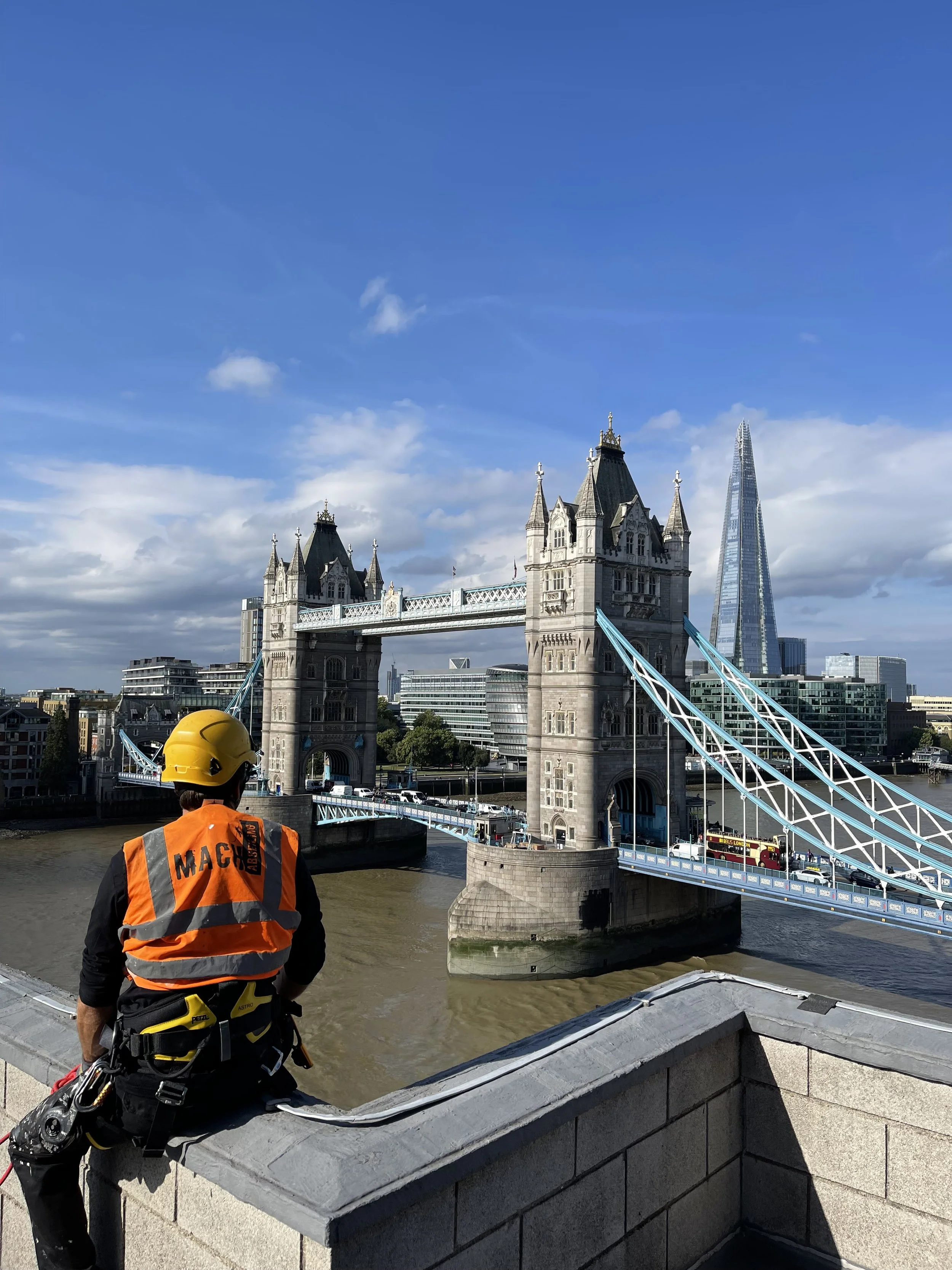 Construction worker with helmet and safety gear sitting on a ledge overlooking Tower Bridge in London with The Shard skyscraper in the background.