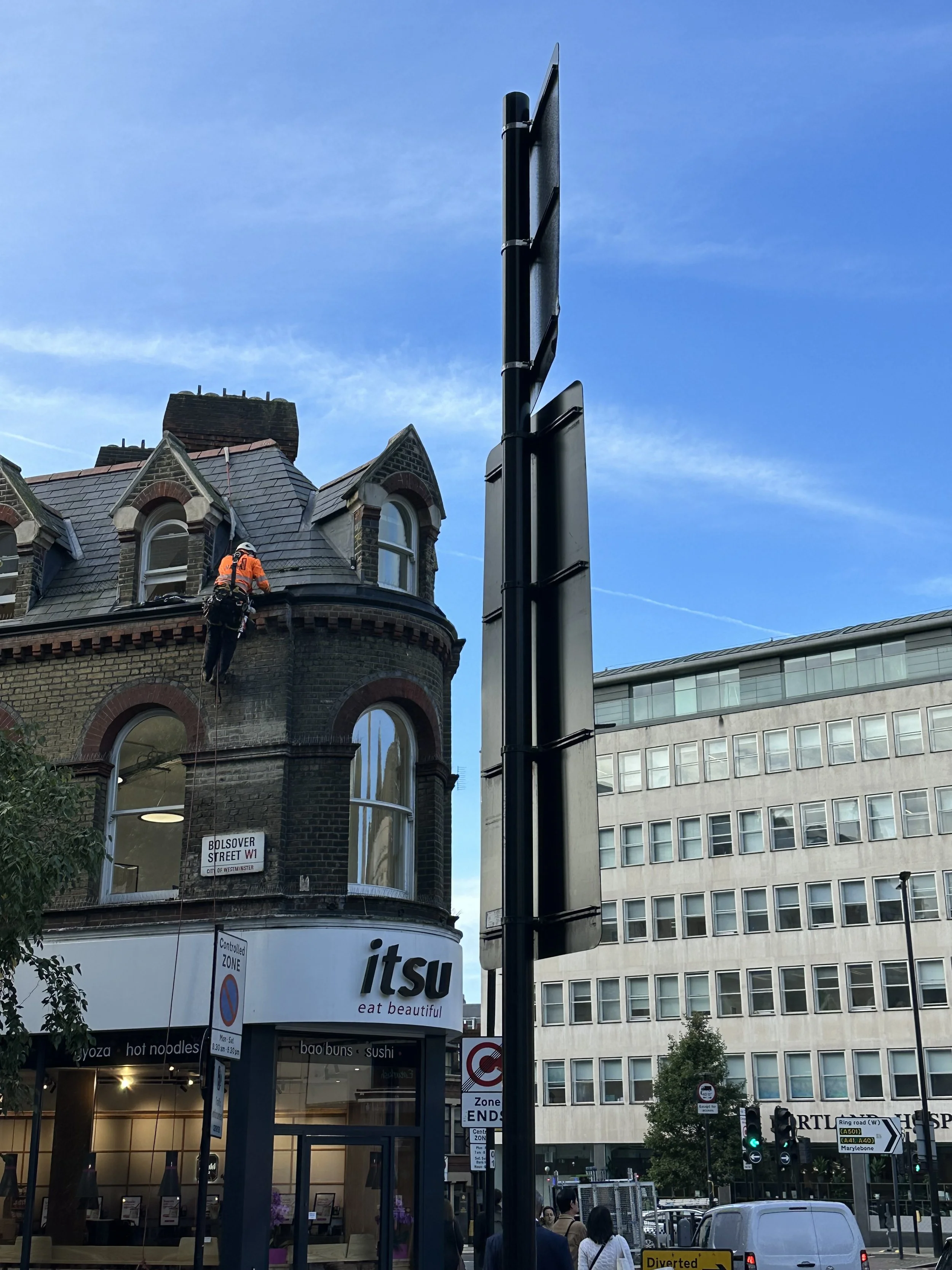 A worker in orange safety gear cleaning the exterior window of a historic brick building with a restaurant called Itsu on the ground floor in an urban street scene. The building is on Bolsover Street in Westminster, London.