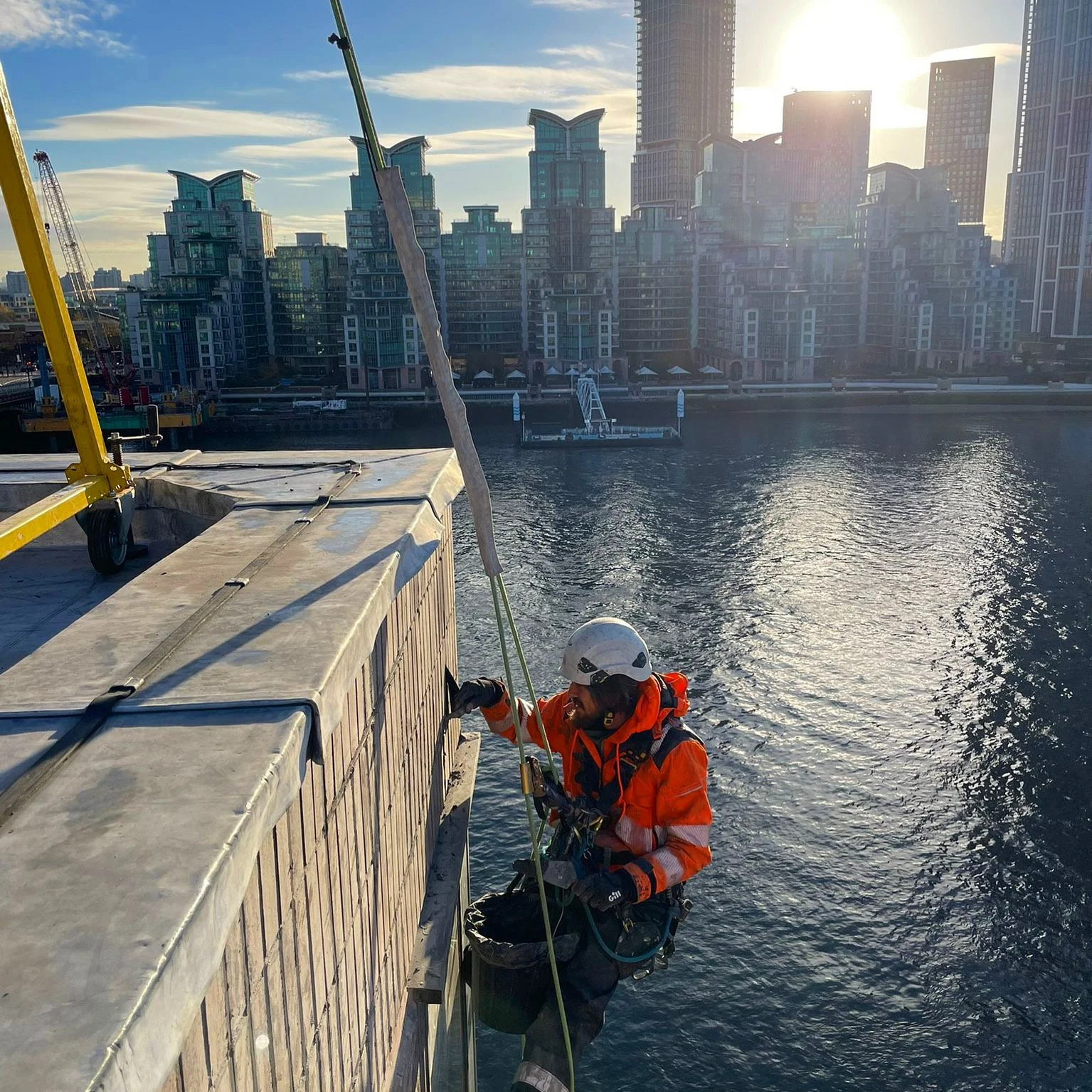 A worker wearing safety gear, including a helmet and harness, is suspended on a rope examining or repairing the exterior of a building beside a body of water with a city skyline in the background.