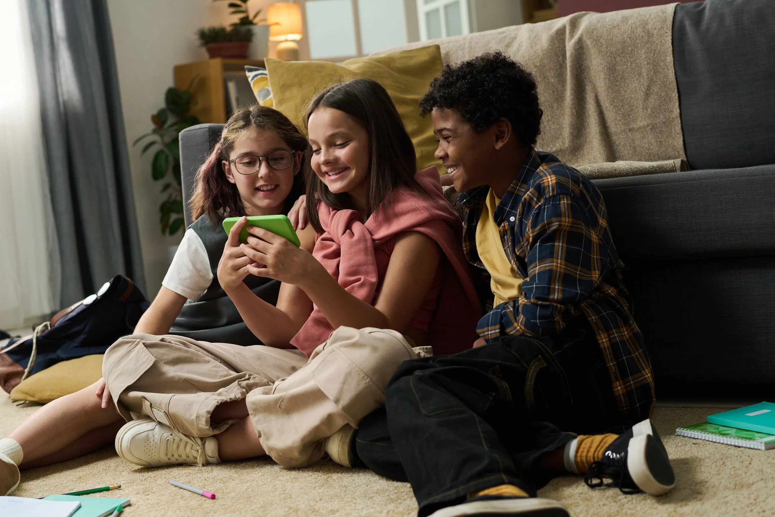 Drie kinderen zitten op de vloer binnen, kijken naar een telefoon en lachen samen.