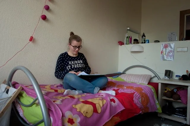 Een jonge vrouw zit op haar bed en leest een boek, in een kamer met kleurrijke dekens en decoraties.