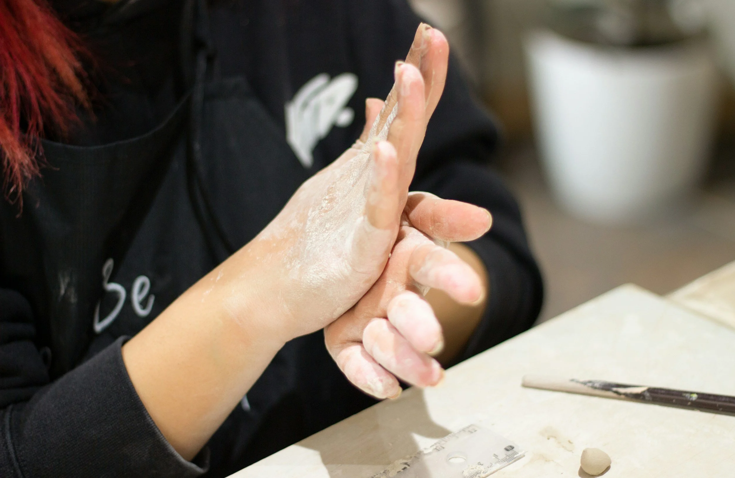 Person rubbing their hands together covered in white flour at a work table with pottery tools and clay.