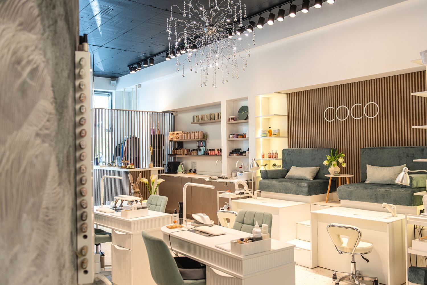 Interior of a modern beauty salon with manicure stations, comfortable seating, and decorative lighting, featuring the word 'COCO' on the back wall.