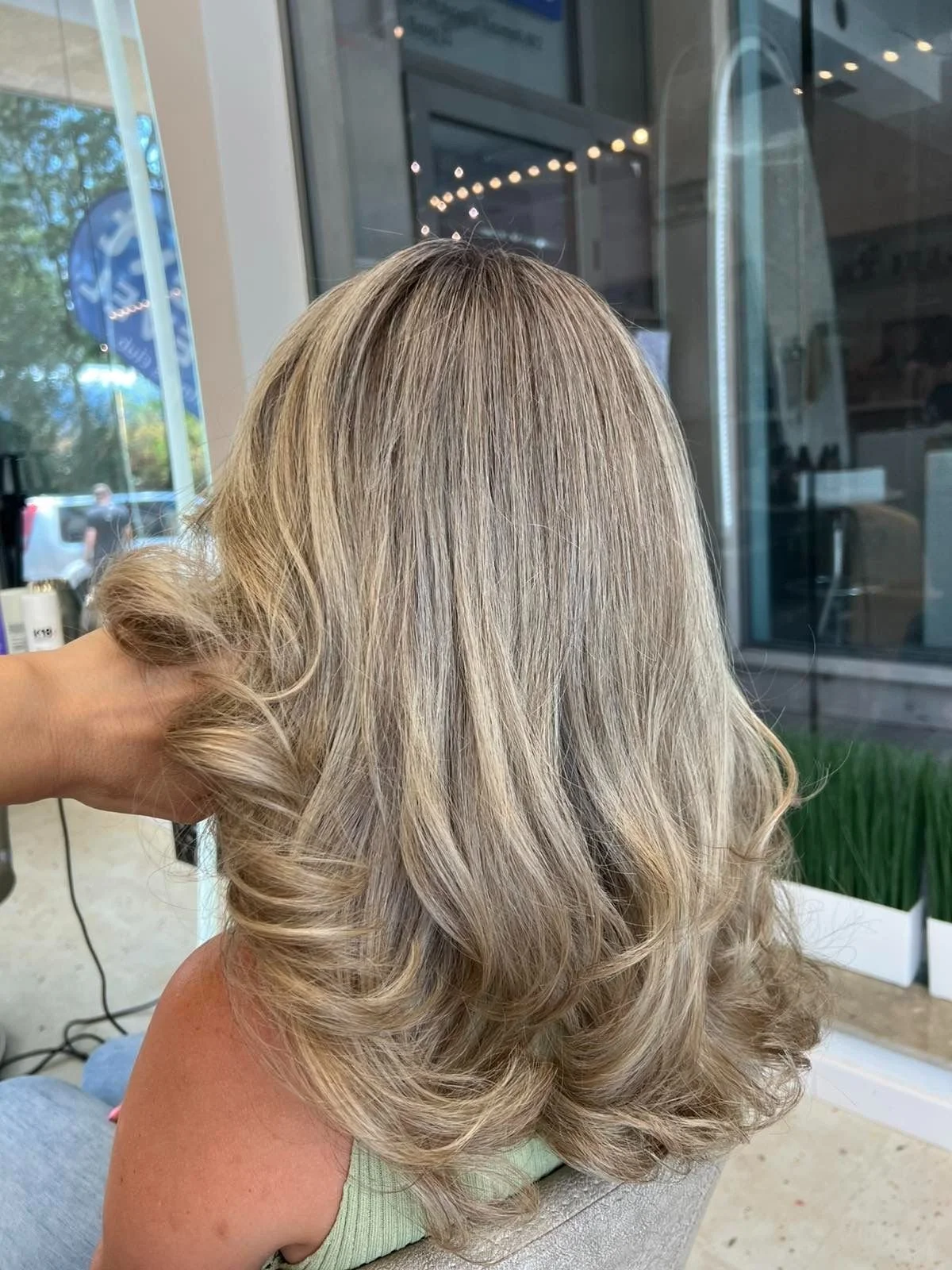 A woman with blonde, styled, wavy hair sitting in a salon near a large window.