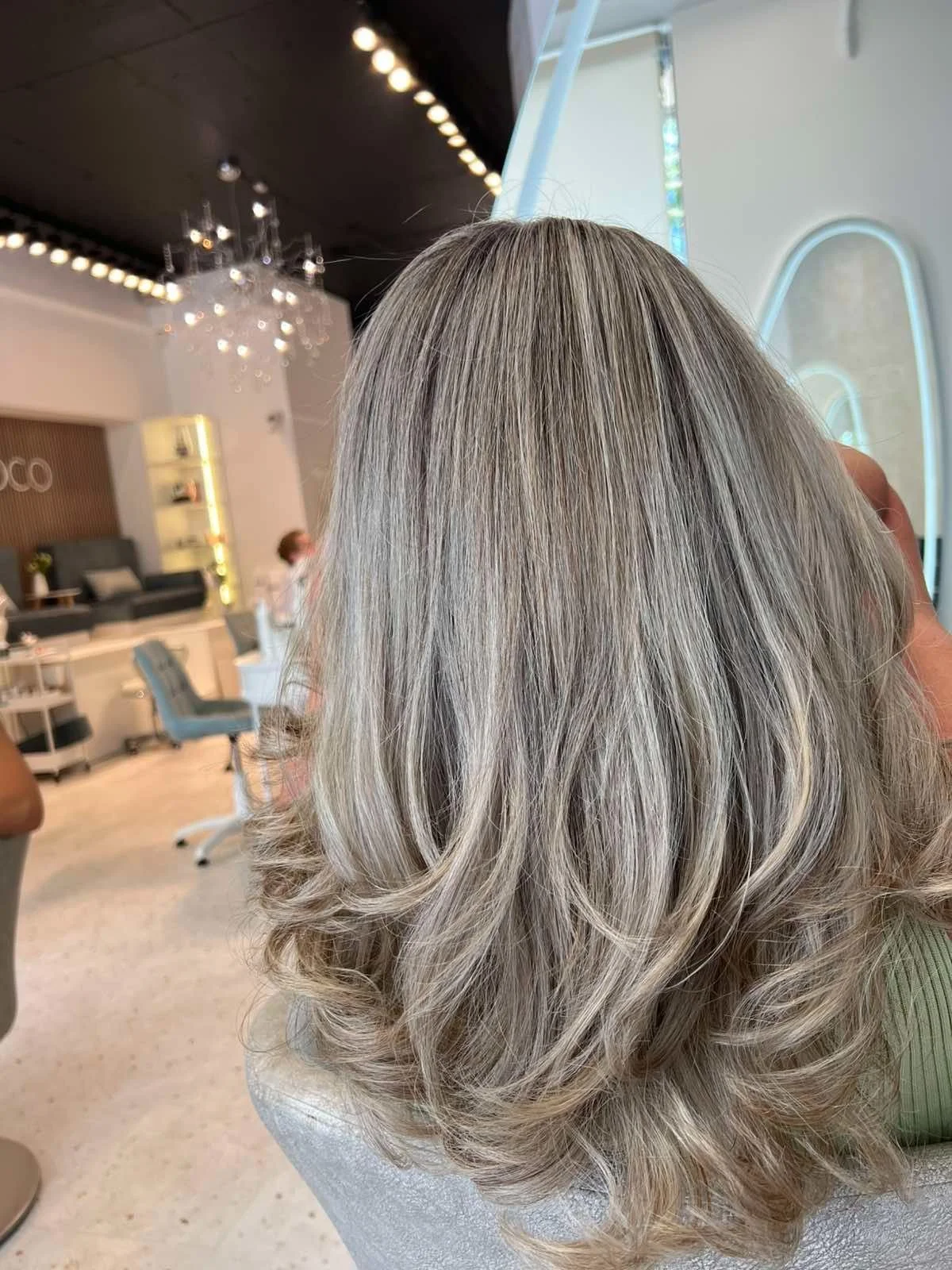 Woman with shoulder-length, wavy, gray-blonde hair sitting in a modern salon.