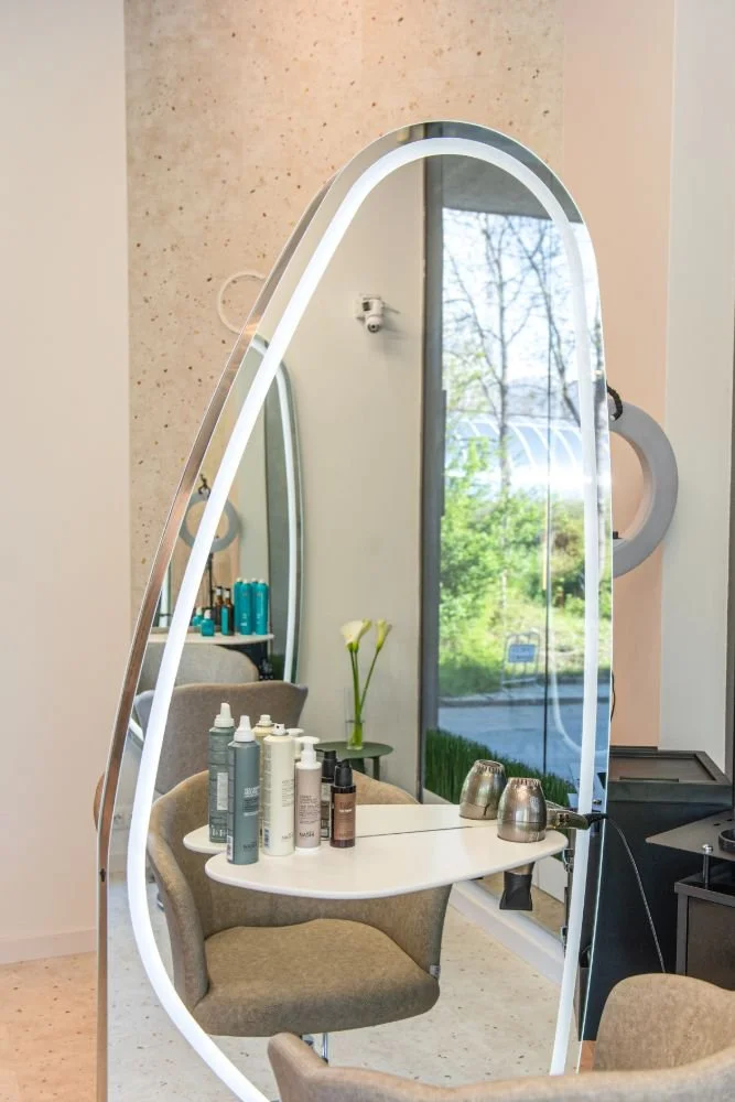 Decorative mirror with LED light outline in a salon, reflecting a table with hair products, a chair, and a vase with two calla lilies, near a large window showing outdoor greenery.
