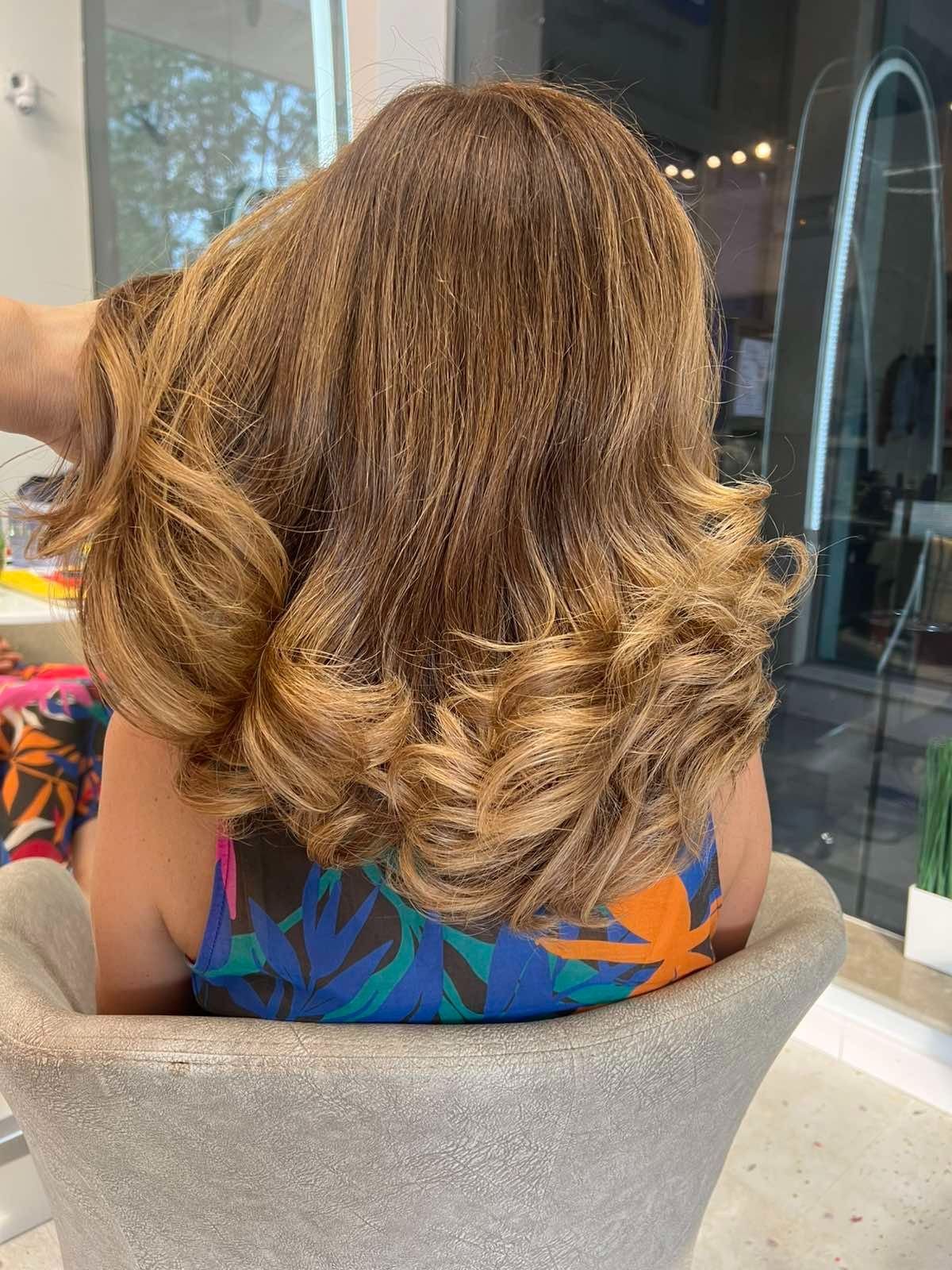 Woman with long, wavy, light brown hair sitting in a salon chair wearing a colorful patterned top.