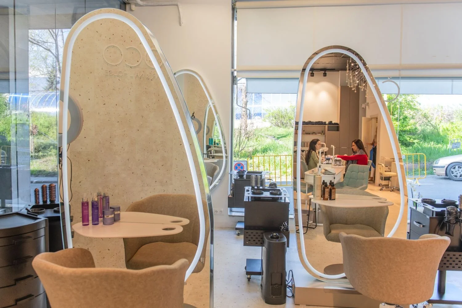 Inside a modern salon with two curved, illuminated partitions and a workspace with chairs, tables, and beauty products. Two women are seated at a table, receiving a manicure or manicure consultation. Large windows reveal a green outdoor area.