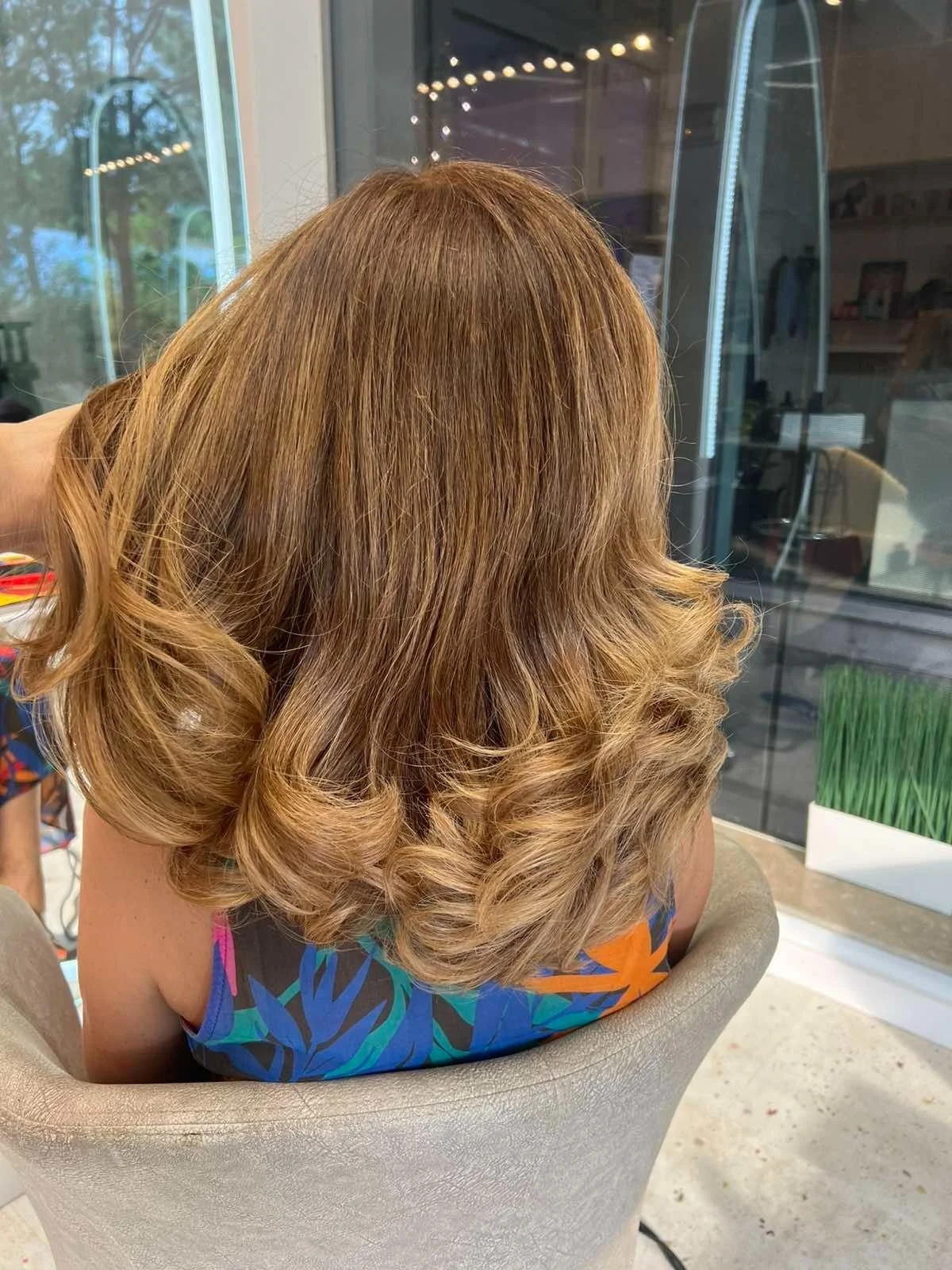 A woman with shoulder-length, wavy, light brown hair, seen from behind, sitting in a salon chair near a large window.