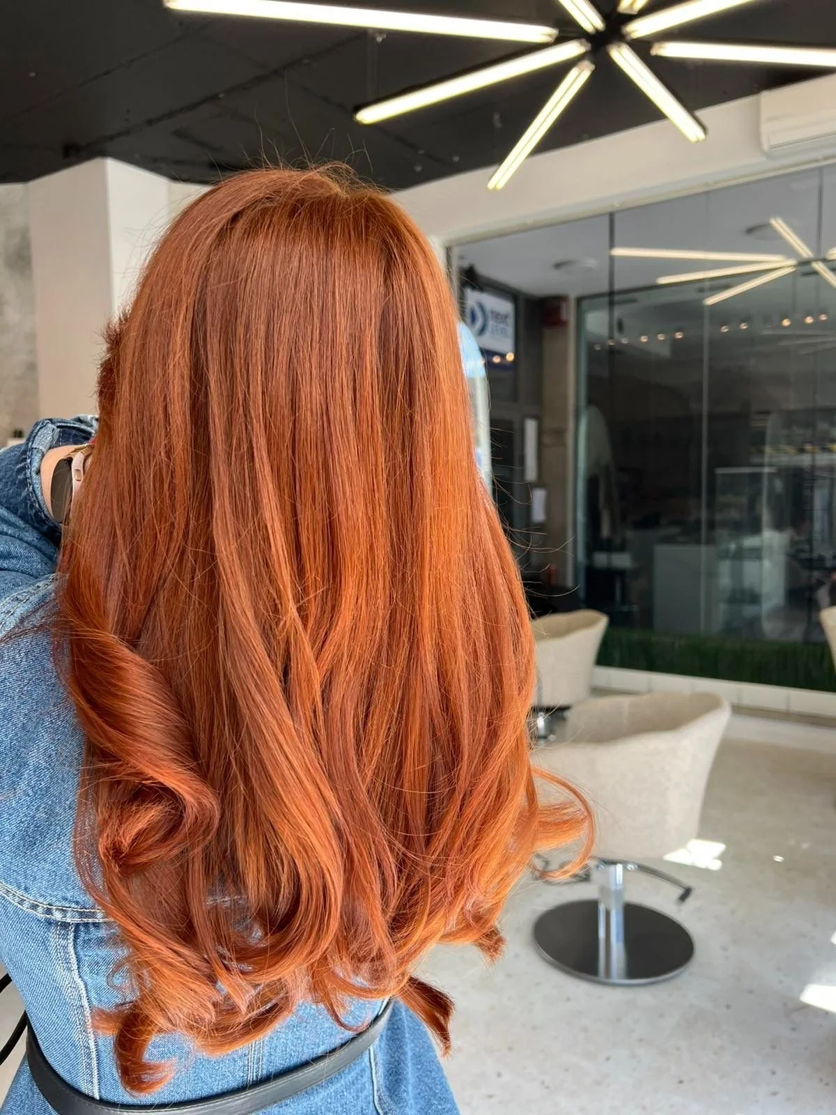 Back of a woman with long, wavy red hair sitting in a modern salon or cafe with contemporary light fixtures and seating.