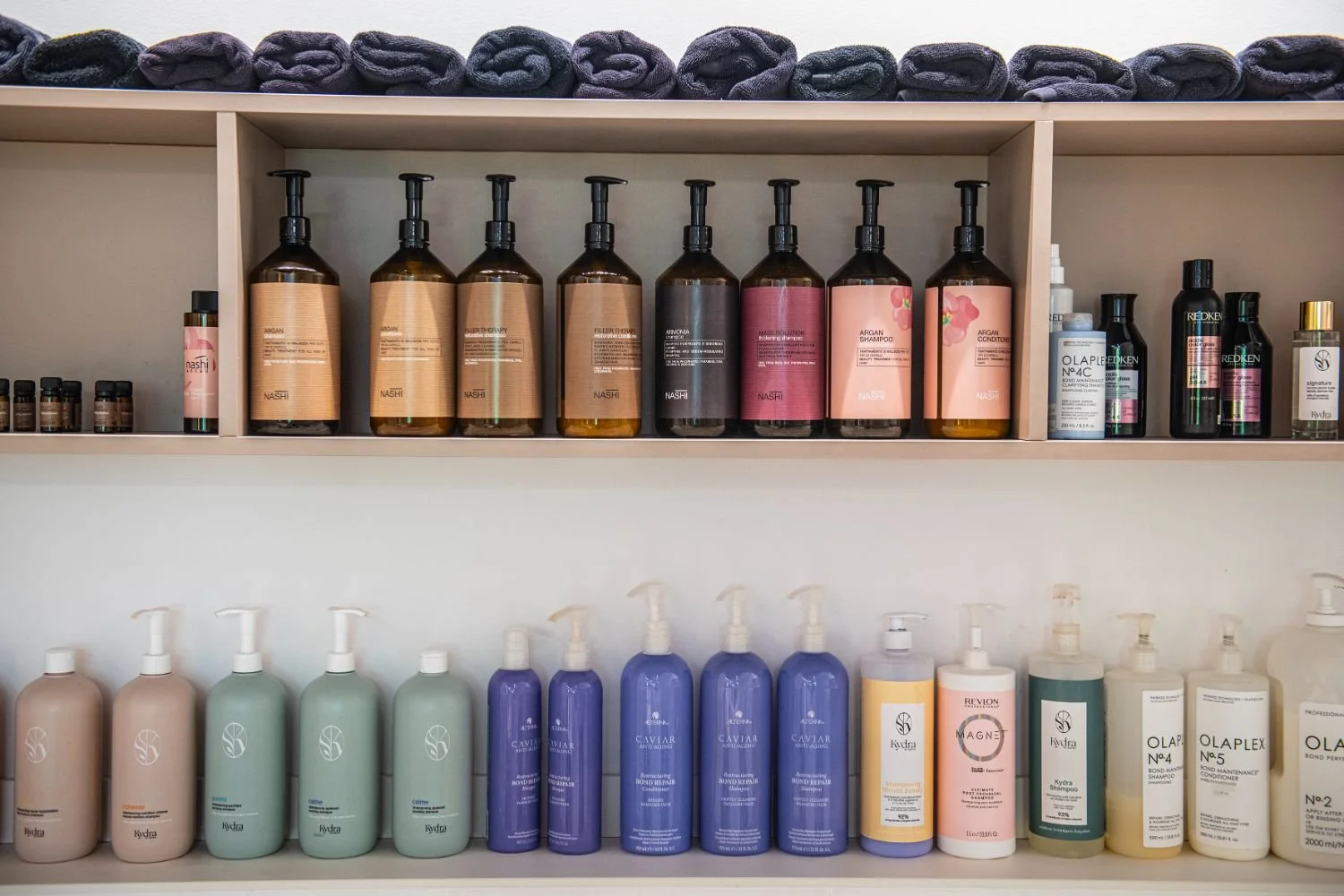 Shelves stocked with various bottles of hair and skincare products, including shampoos, conditioners, and serums, arranged neatly in a salon or spa setting.
