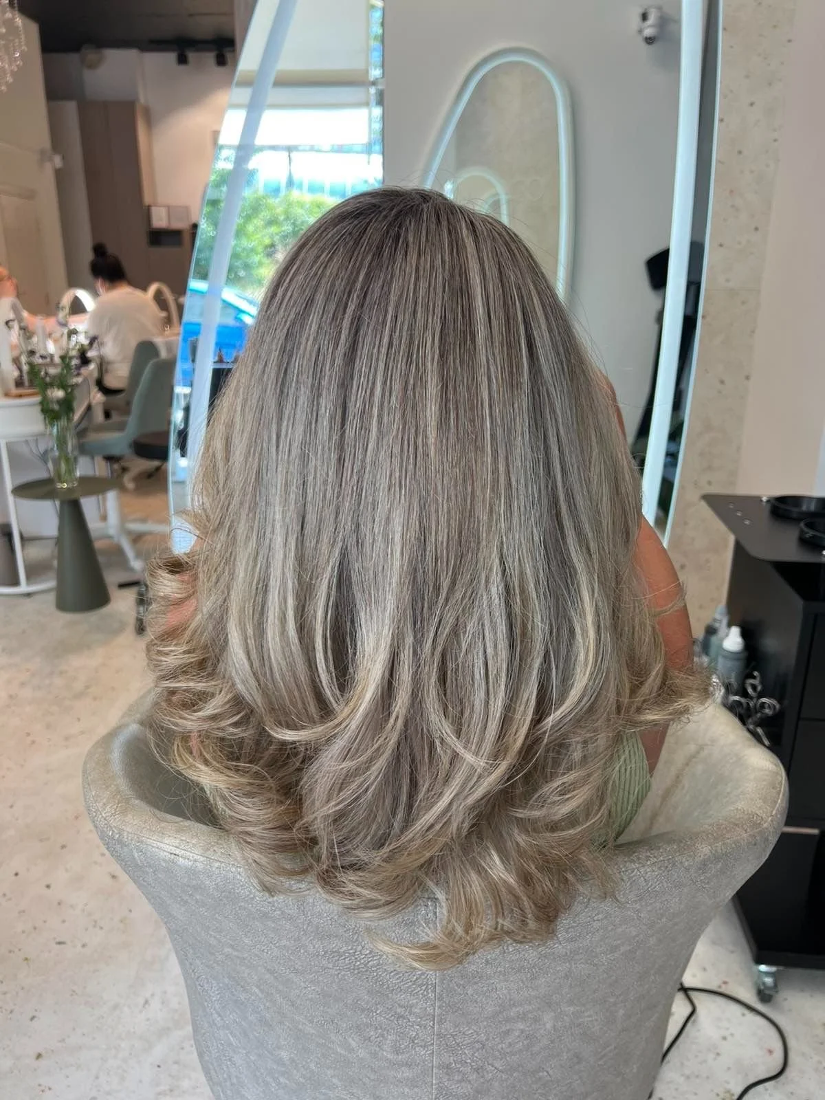 Back of a woman's head with shoulder-length, styled, blonde hair showing shades of silver and gray, seated in a salon chair.