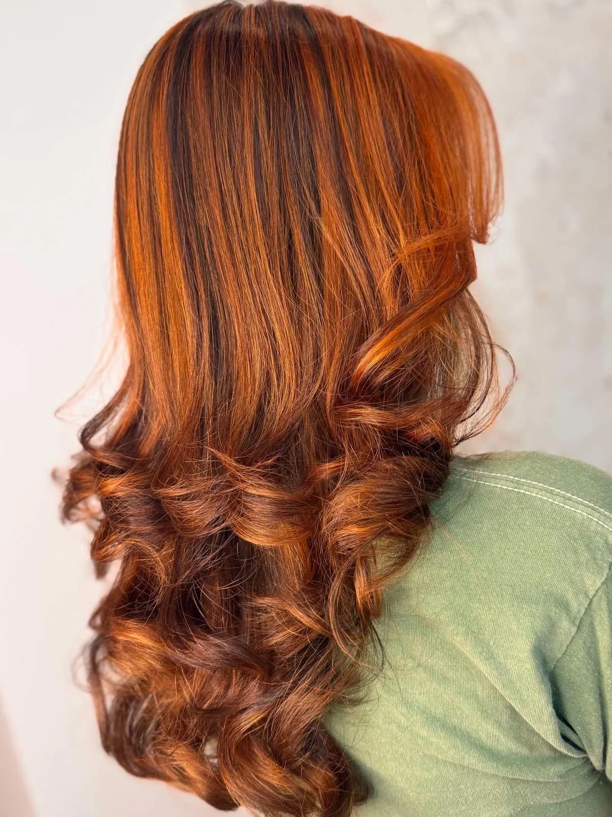 A close-up of a woman's head showing long, wavy, red-orange hair with darker roots, falling over her shoulder against a plain background, with part of a green shirt visible.