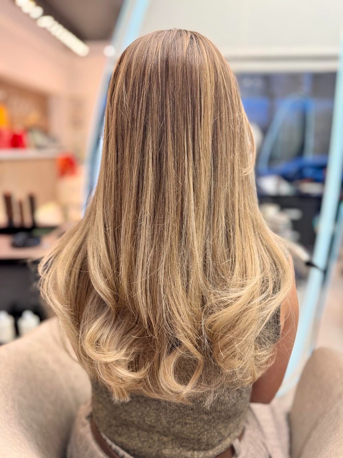 Back view of a woman with long, blonde, wavy hair sitting in a salon.