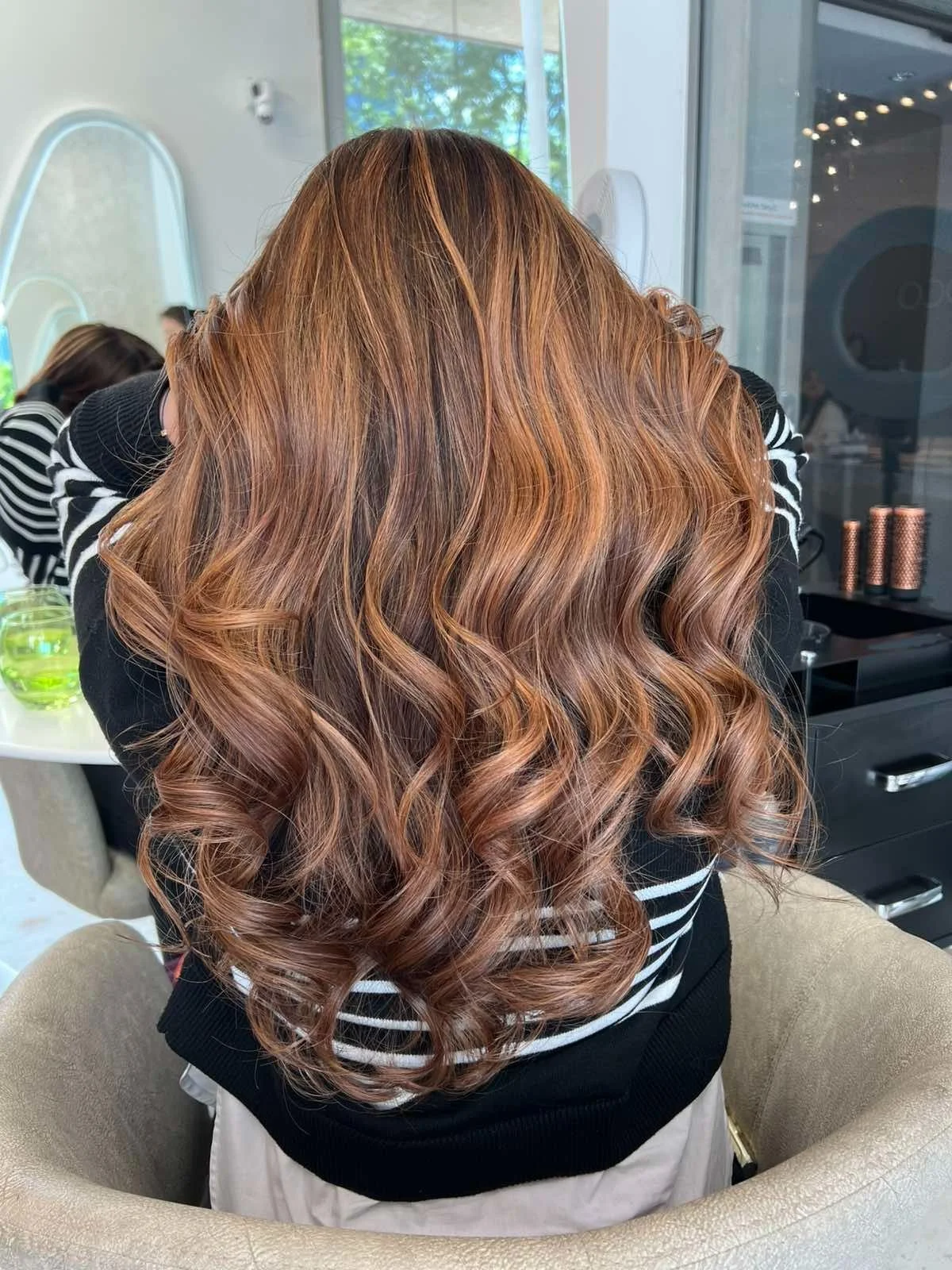Back view of a woman with long, styled, wavy auburn hair sitting in a salon chair.